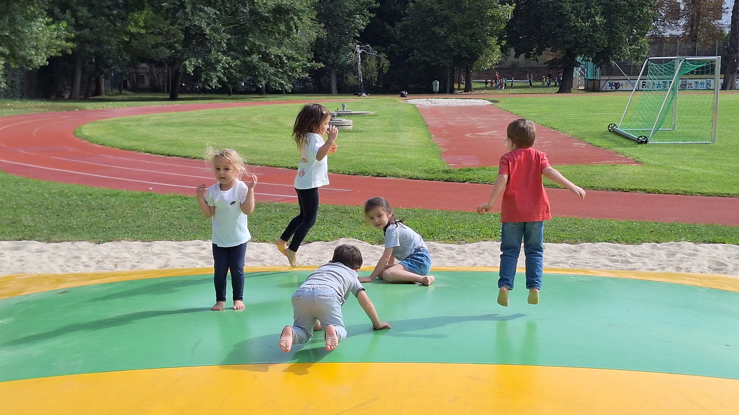 Fünf kleine Kinder auf einer Hüpfblase auf einem Sportplatz mit Leichtathletikbahn in sommerlicher Kleidung hüpfend und knieend.