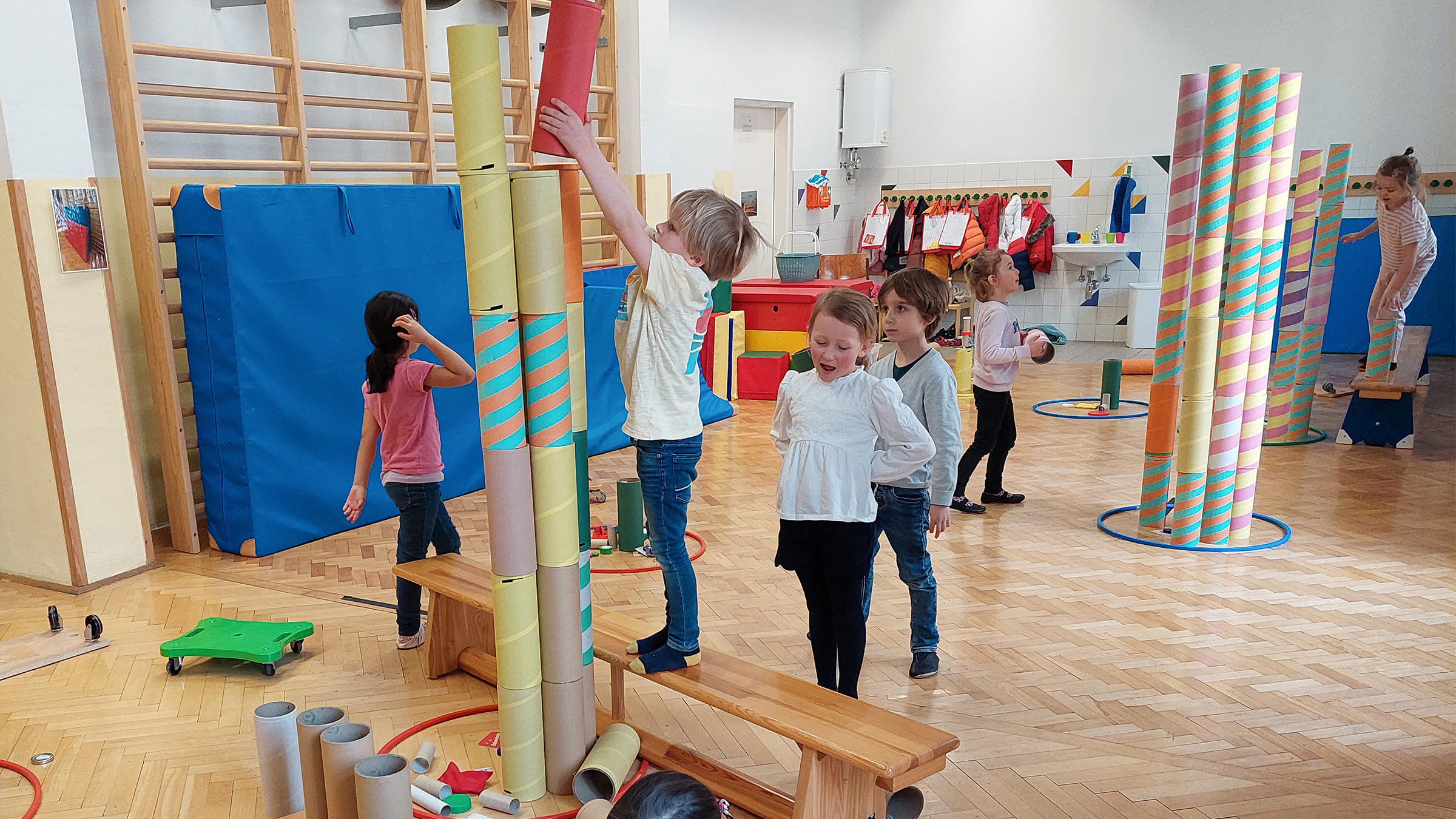 Gruppe von Kindern in einem Turnsaal, die mit bunten Pappröhren hohe Türme bauen und dazu auf Langbänke steigen.