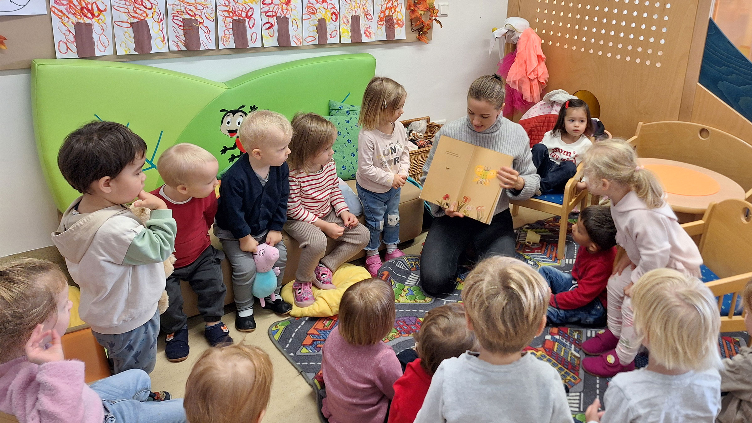 Gruppe von Kleinkindern in einem Kreis am Boden sitzend und zu einer erwachsenen Frau hin blickend, die ein aufgeklapptes Buch erklärt