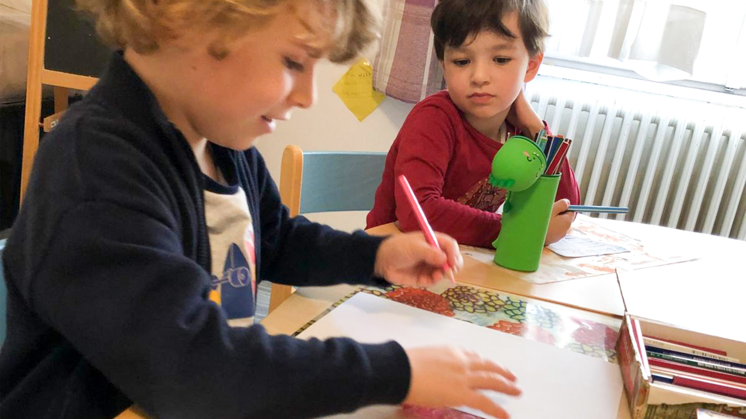 Zwei kleine Buben an einem Tisch sitzend, einer von ihnen konzentriert sich auf den Stift in seiner Hand, der andere sieht ihm zu.
