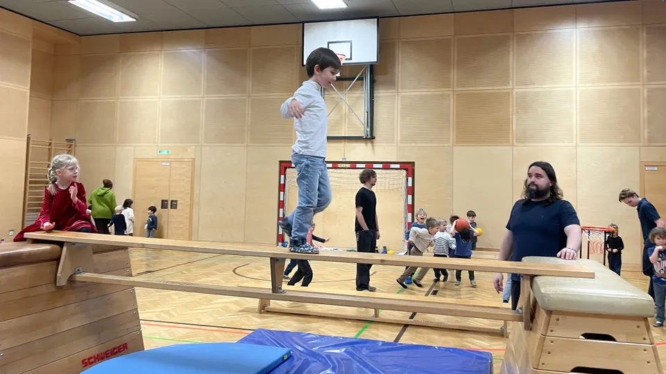 Ein Bub in einem Turnsaal auf einer Langbank wie auf einer Brücke mit ausgestreckten Händen darüber balancierend mit einem erwachsenen Mann mit Vollbart daneben stehend und vielen Kindern im Hintergrund.