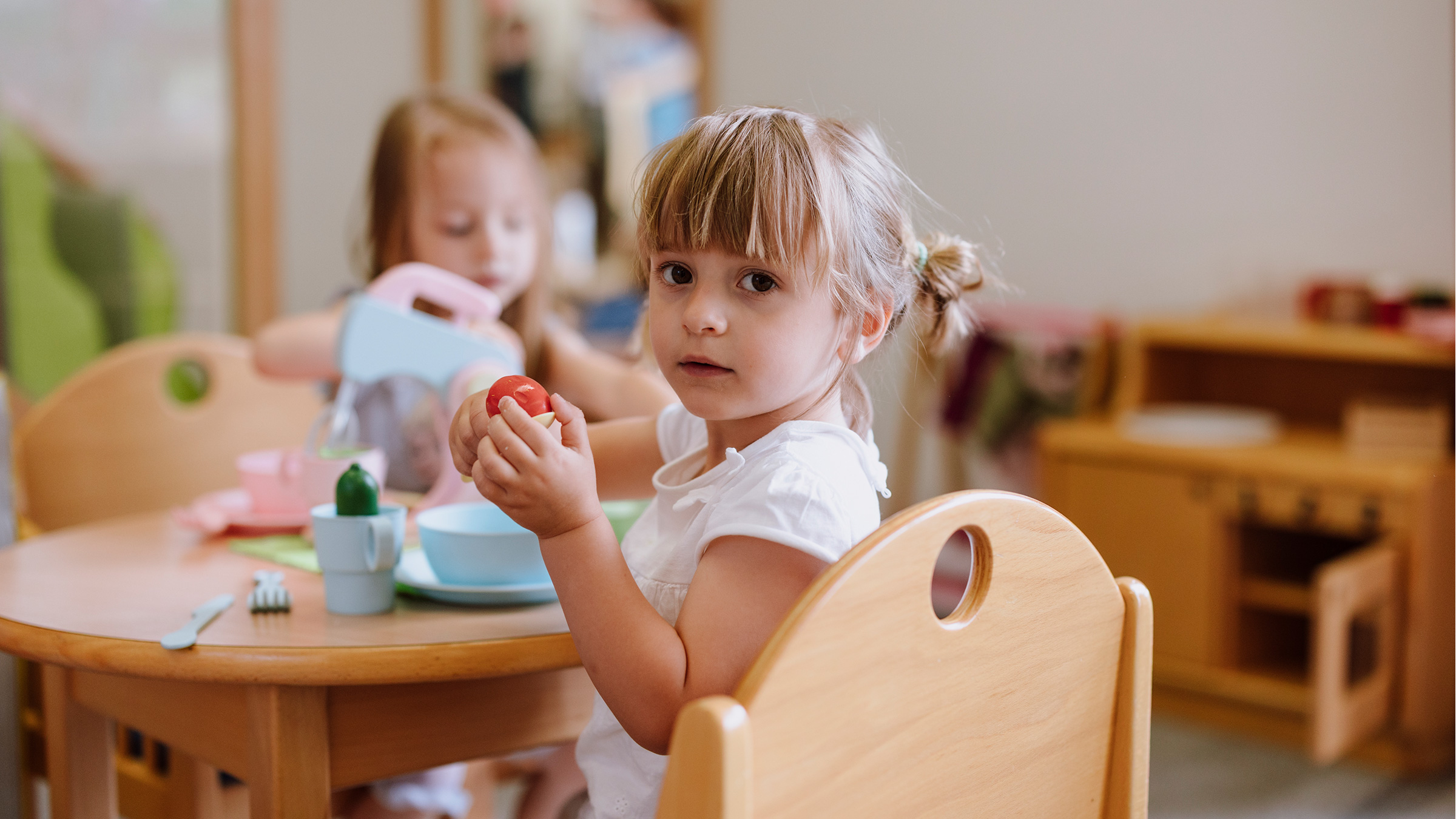 Zwei kleine Mädchen an einem Kindertisch sitzend mit Kindergeschirr und Mixer spielend. Ein Mädchen dreht den Kopf zur Kamera