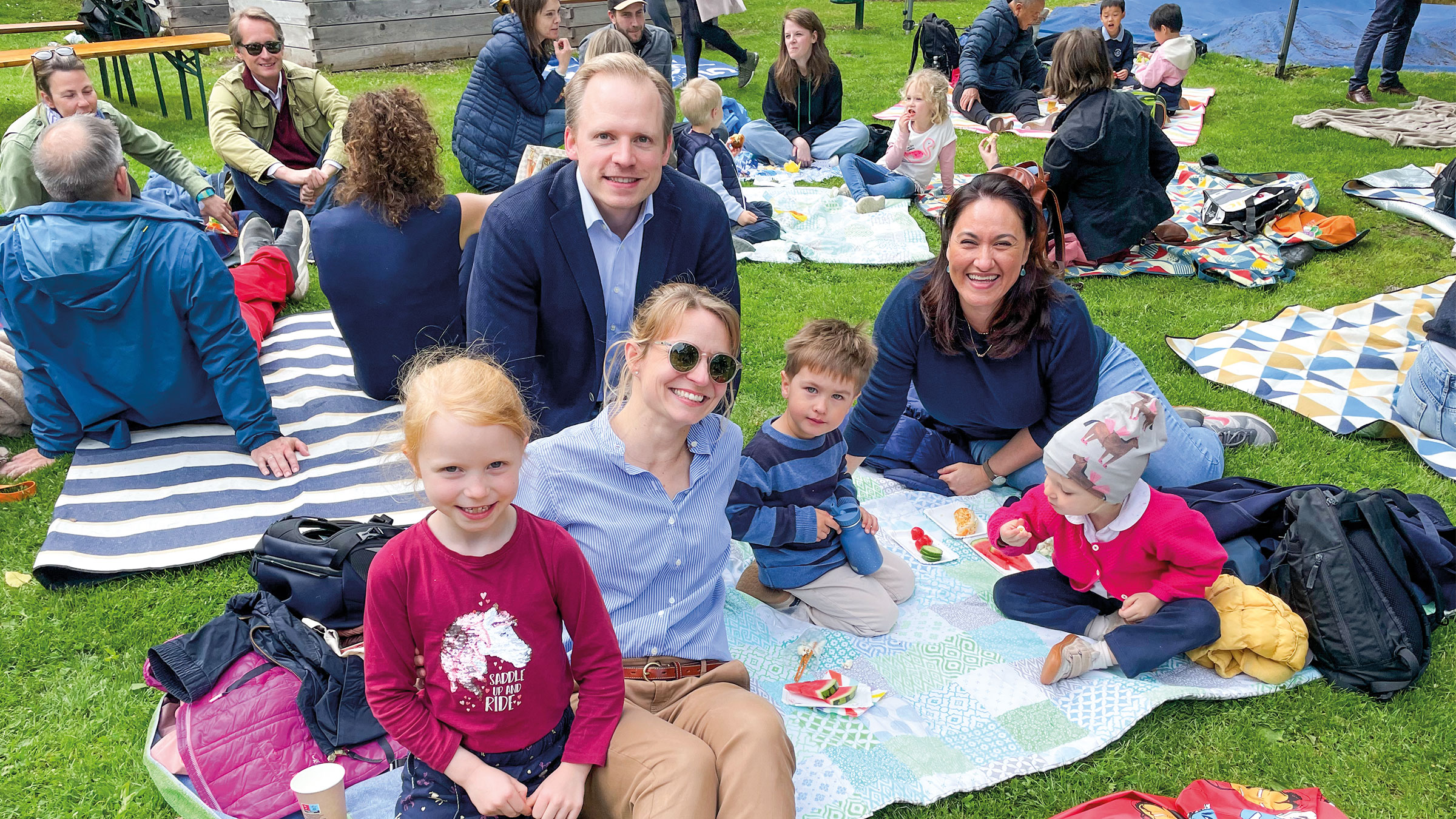 Erwachsene Menschen und Kinder verschiedenen Alters gemütlich und gut gelaunt auf Picknickdecken auf einer Wiese sitzend