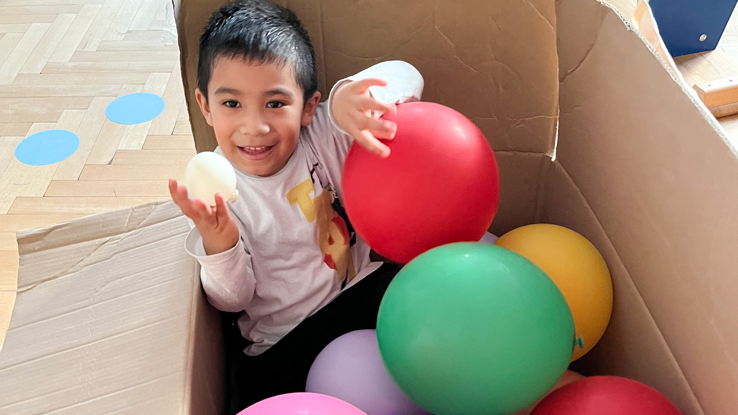 Bub mit dunklen Haaren in einem großen Karton mit bunten Luftballons sitzend und lachend einen kleinen weißen Ballon haltend.