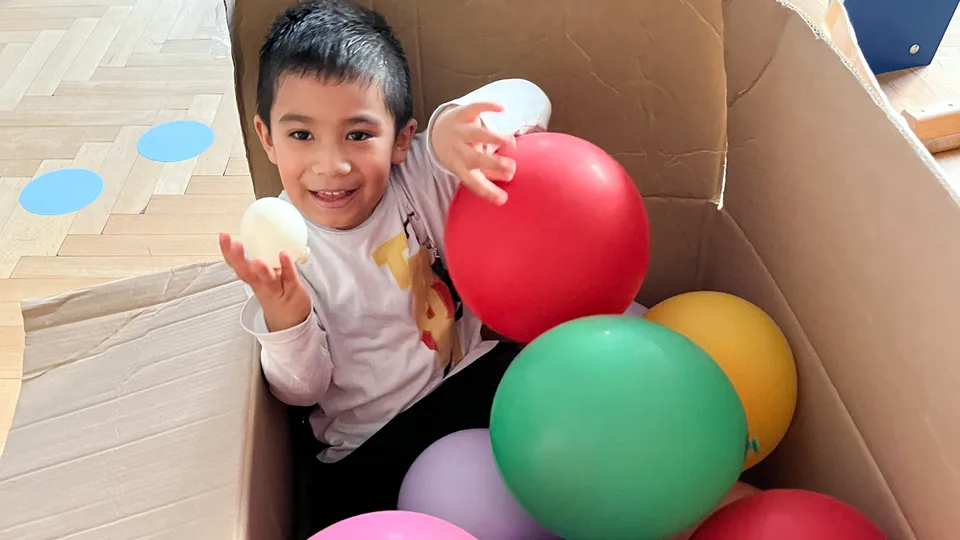 Bub mit dunklen Haaren in einem großen Karton mit bunten Luftballons sitzend und lachend einen kleinen weißen Ballon haltend.