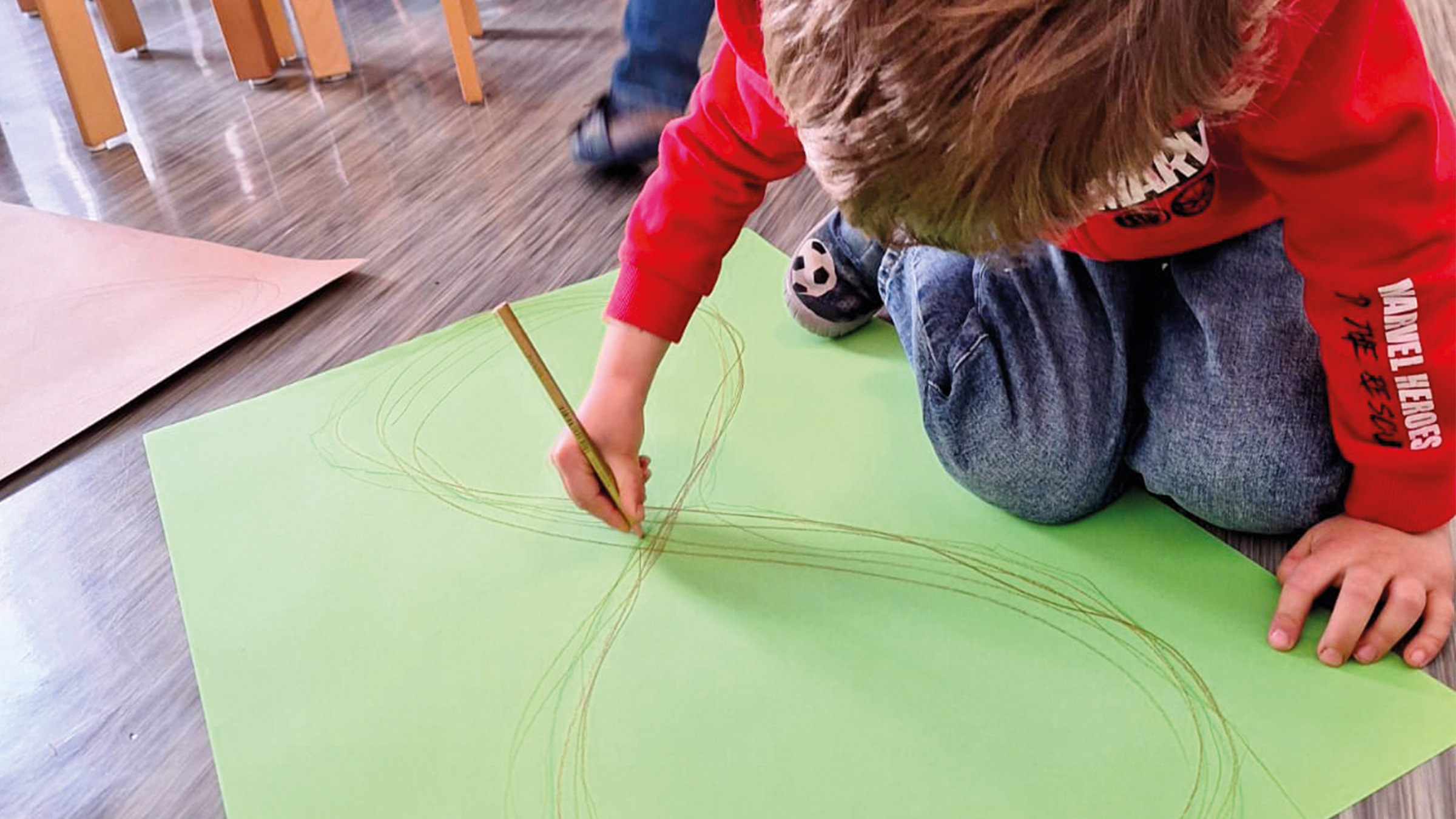 Bub mit rotem Pulli und Jeans am Boden sitzend und mit einem Stift auf einem großen grünen Bogen Papier einen Achter zeichnend.