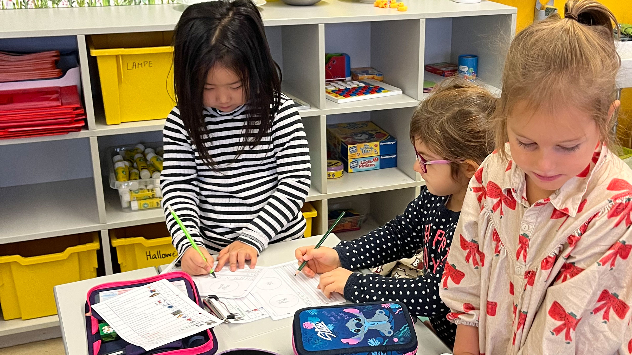 Drei kleine Mädchen in einem Klassenzimmer auf Zetteln schreibend.