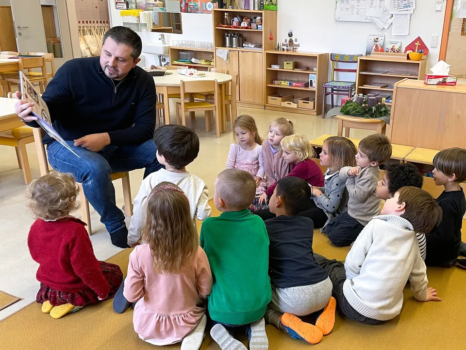 Erwachsender Mann auf einem Stuhl in einem Kindergarten sitzend mit einer Gruppe von kleinen Kindern im Halbkreis um ihn herum sitzend und auf das Buch in seiner Hand blickend.