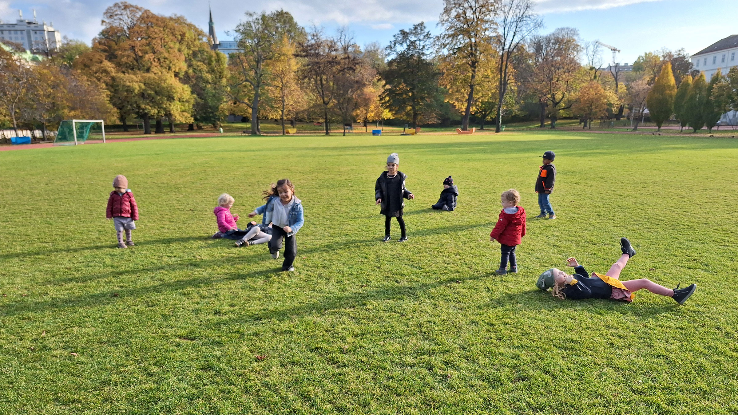 Kleinere Kinder auf einer großen Wiese mit Bäumen im Hintergrund auf der Wiese tobend und laufend.