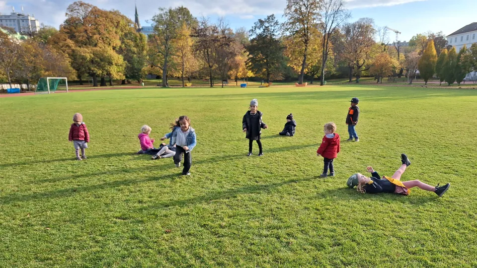 Kleinere Kinder auf einer großen Wiese mit Bäumen im Hintergrund auf der Wiese tobend und laufend.
