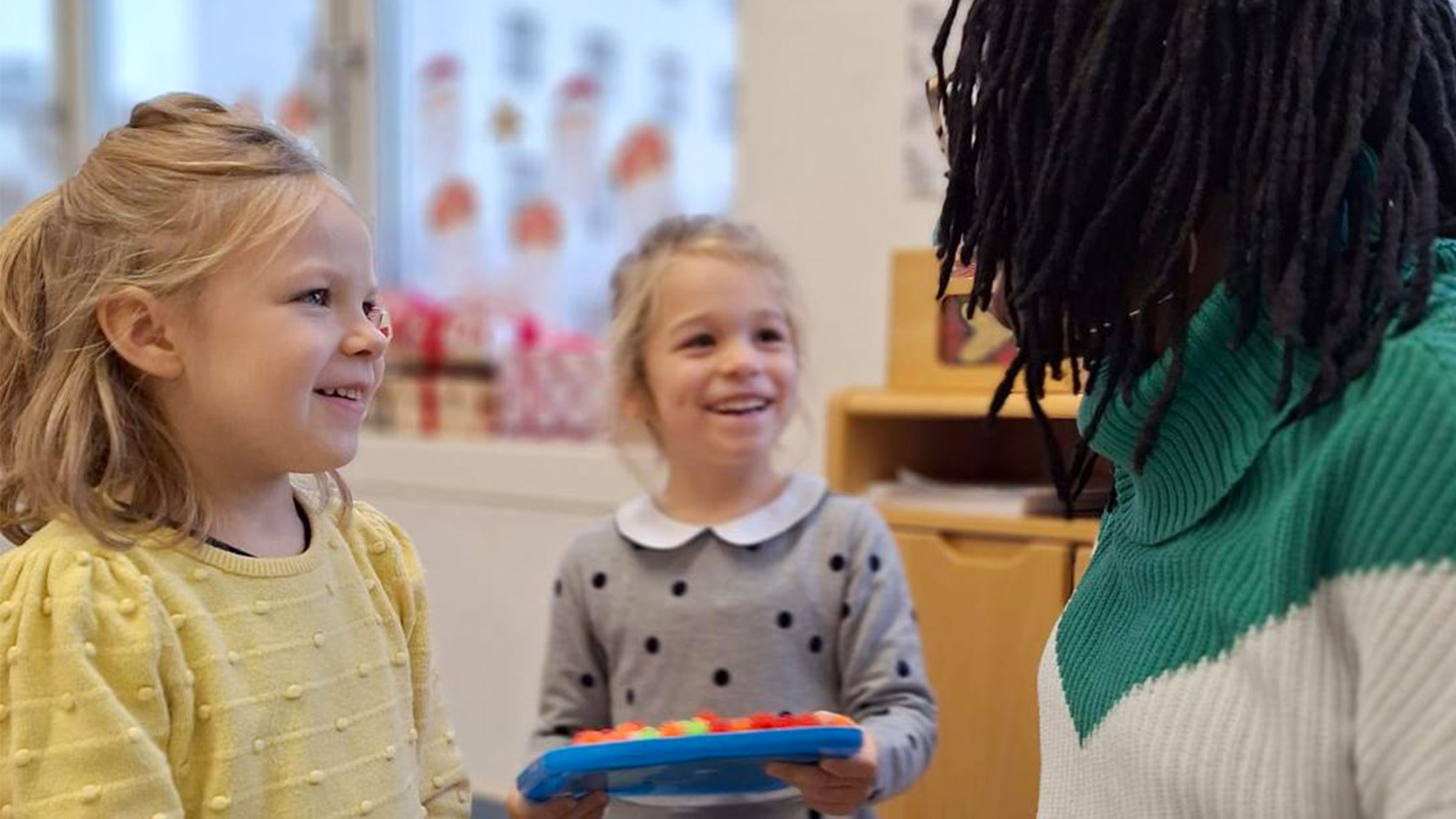 Zwei blonde Mädchen eine Frau mit weggedrehtem Gesicht und schwarzen Dreadlocks anstrahlend.