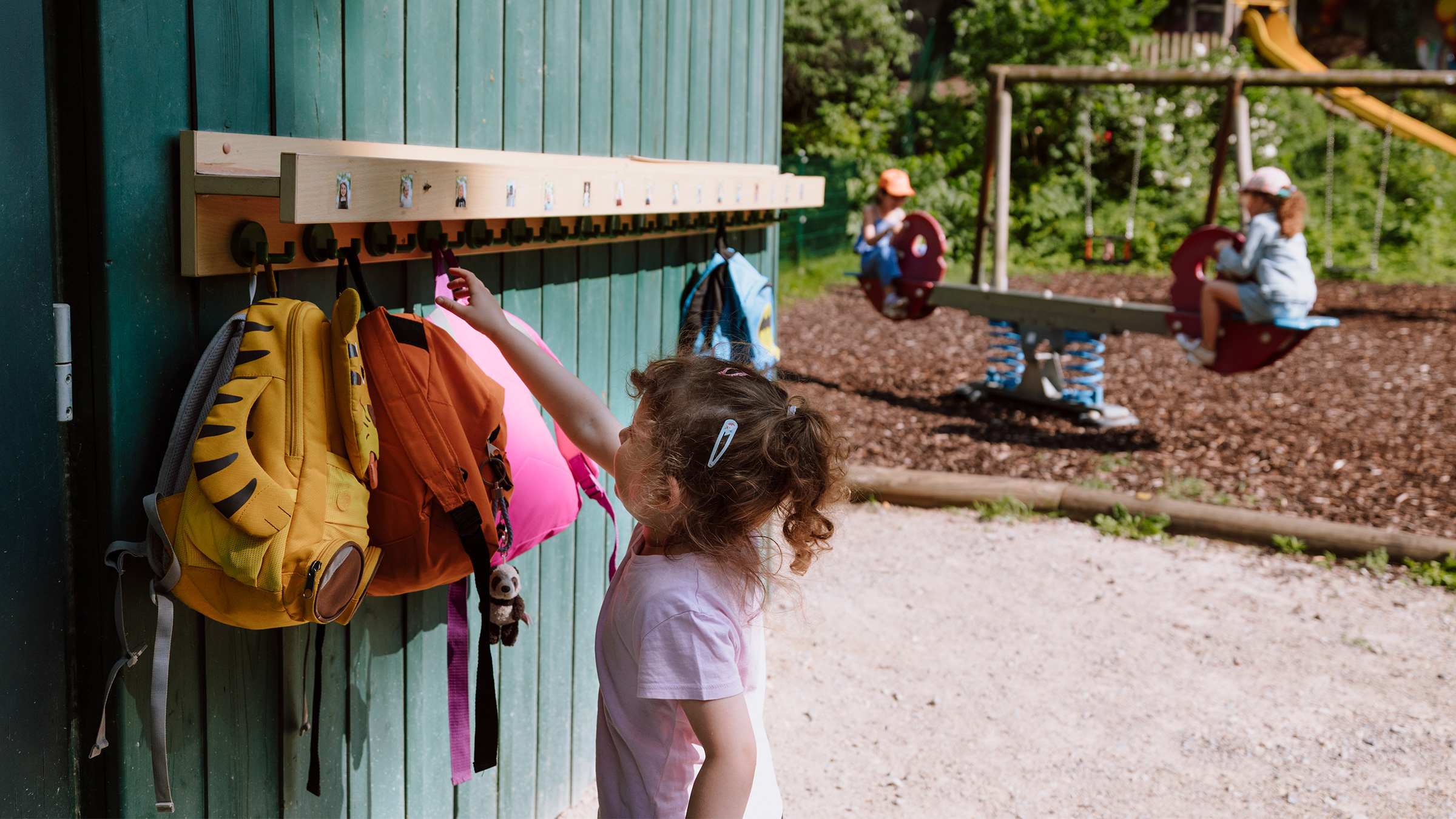 Mädchen mit lockigem Pferdeschwanz an einem sonnigen Tag auf einem Spielplatz einen Rucksack an einen Garderobenhaken hängend mit zwei weiteren Mädchen im Hintergrund auf einer Wippe sitzend.