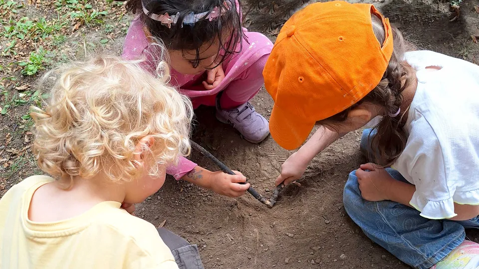 Drei kleine Kinder mit Stöckchen in der Erde grabend, eines von ihnen mit blonden Locken, ein Mädchen mit dunklen Haaren und Haarreifen, eines mit oranger Kappe