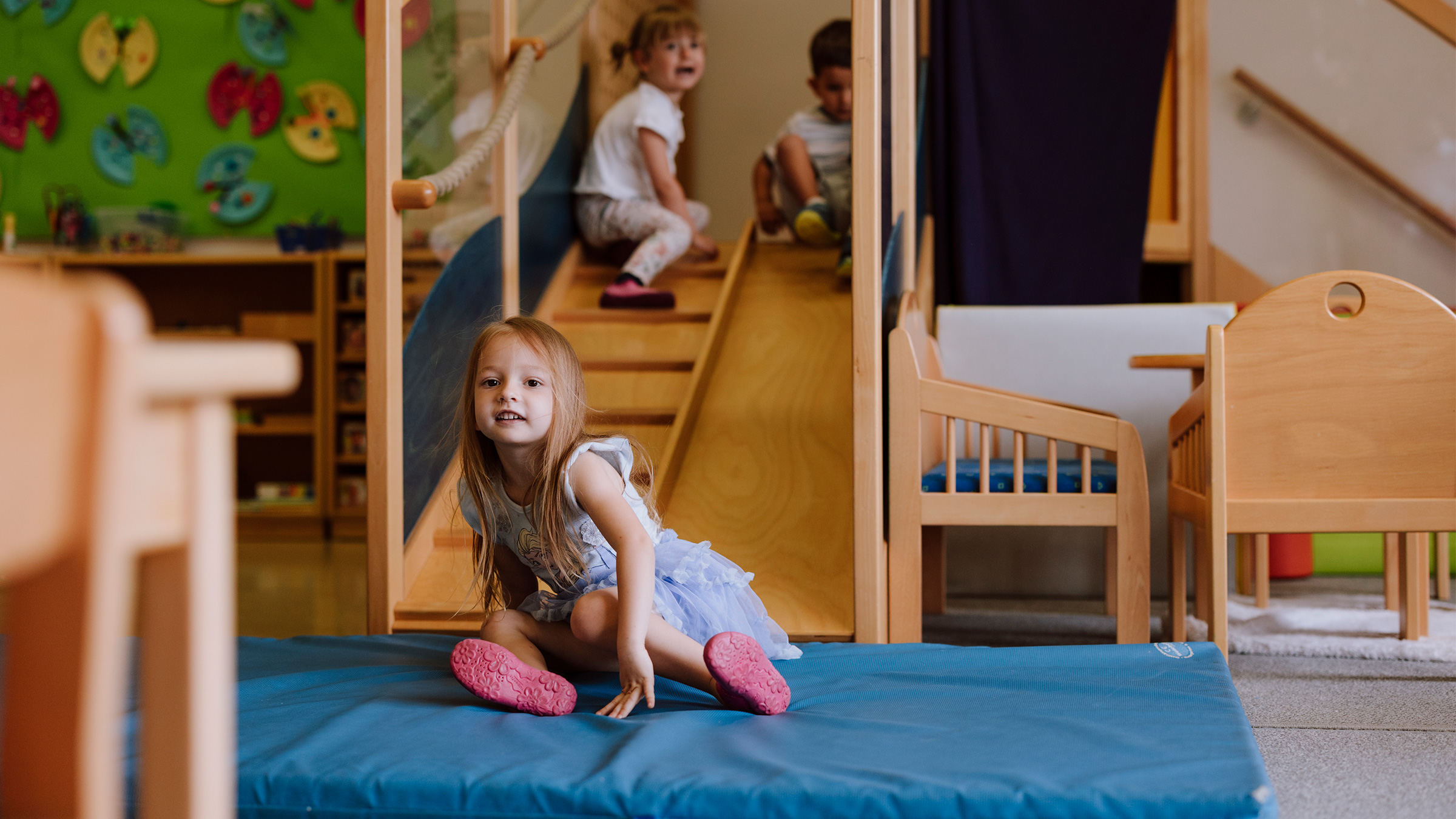 Drei kleine Kinder in einem Kindergarten auf einer Rutsche im Raum auf eine blaue Matte hinunter rutschend.