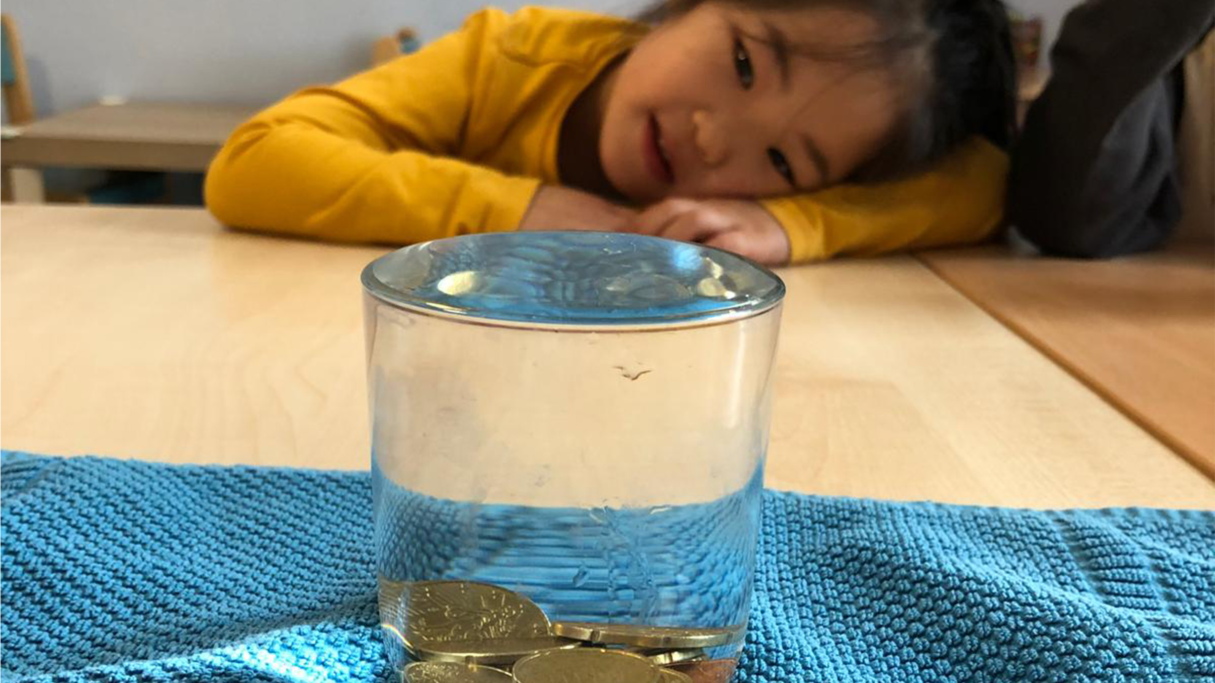 Ein Glas randvoll mit Wasser und Münzen drinnen auf einem blauen Handtuch stehend mit einem Mädchen mit gelbem Pullover im Hintergrund und seitlich am Tisch liegenden Kopf fasziniert das Glas betrachend.