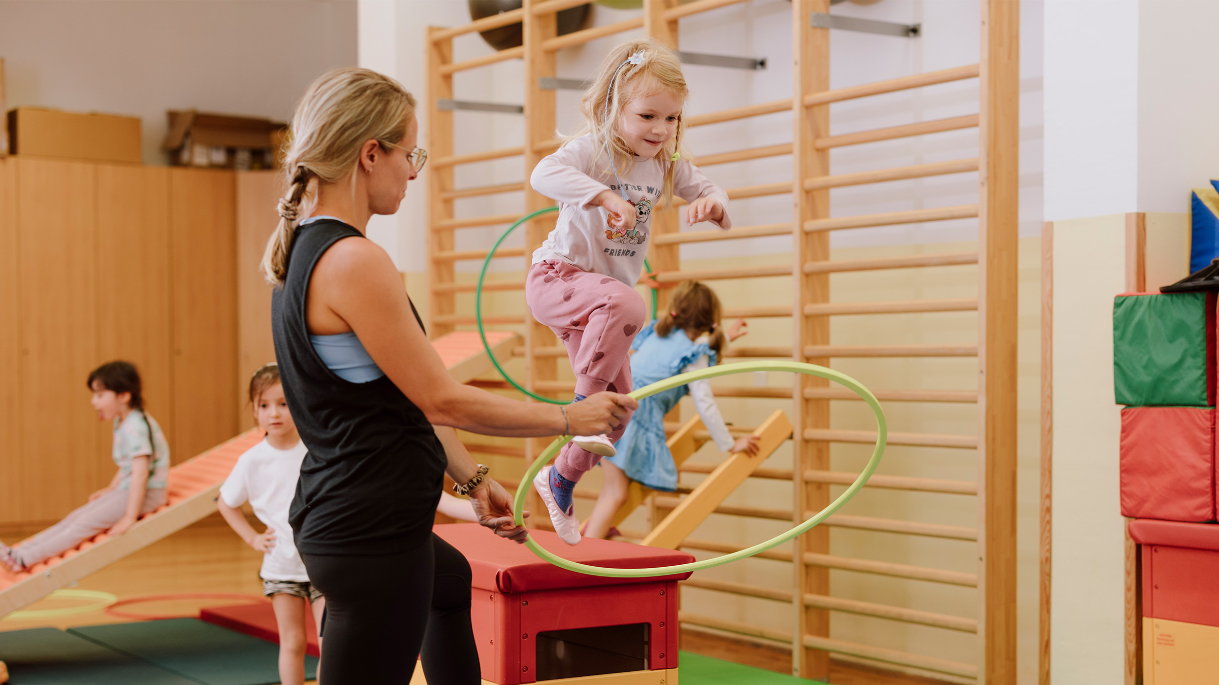 Kleines Mädchen von einem Kasten im Turnsaal durch einen hellgrünen Ring, von der Trainerin gehalten, auf einen Weichboden springend.