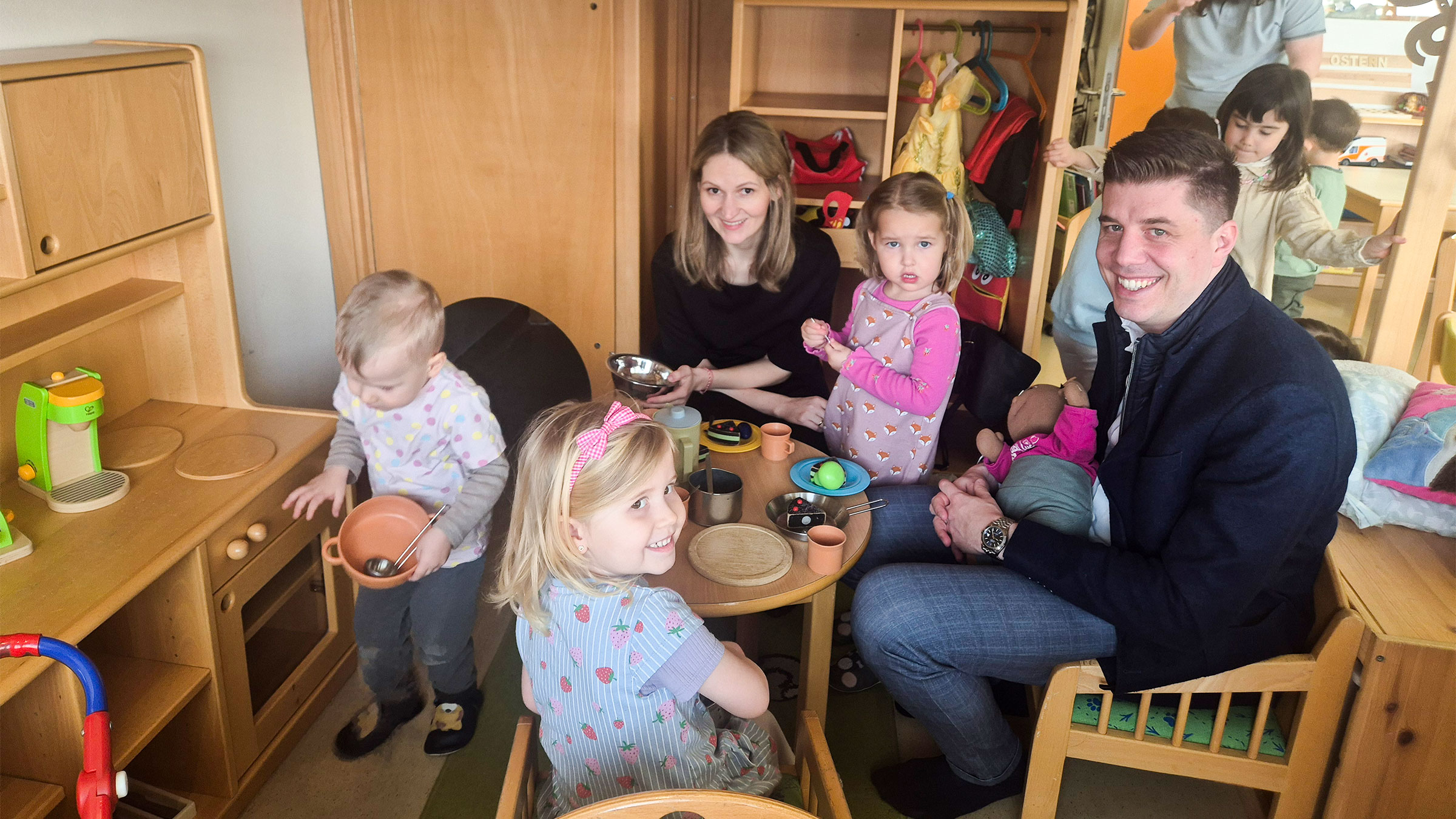 Ein Mann und eine Frau in einer Kinderecke an einem Kindertisch mit drei kleinen Mädchen sitzend und die Kamera lachend.