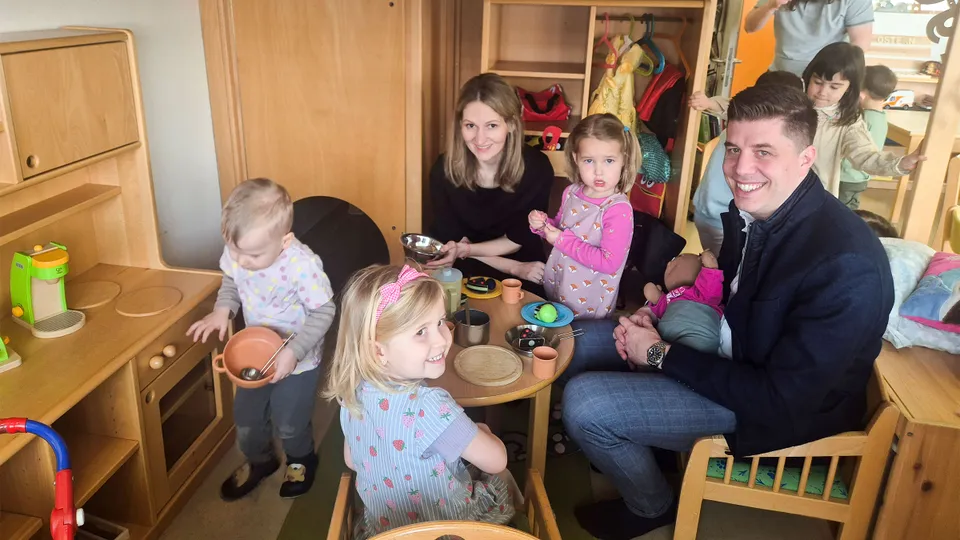 Ein Mann und eine Frau in einer Kinderecke an einem Kindertisch mit drei kleinen Mädchen sitzend und die Kamera lachend.