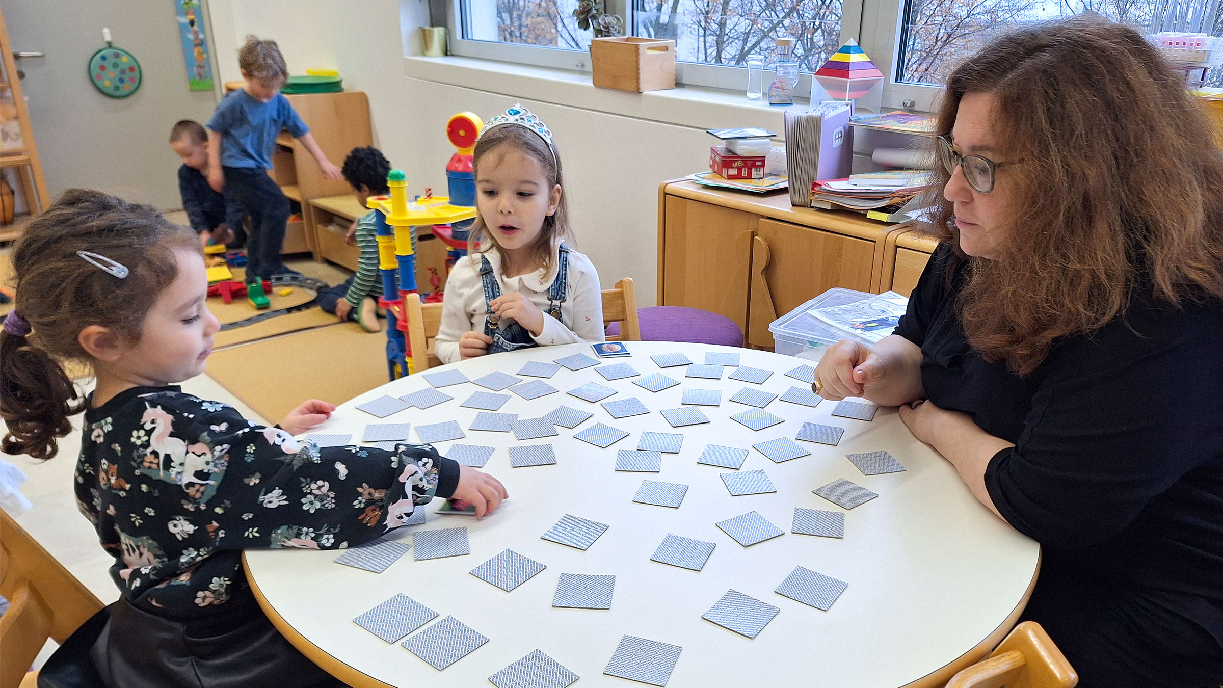 Zwei kleine Mädchen mit einer Frau an einem Kindertisch sitzend und Memory spielend