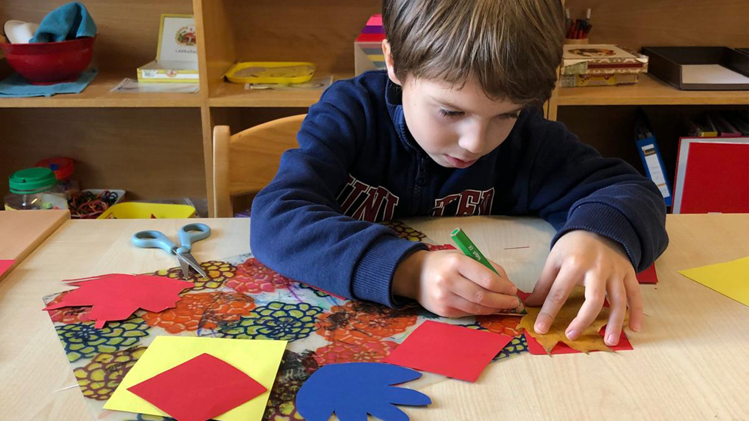 Ein Bub der konzentriert mit einem grünen Buntstift die Ränder eines Blattes markiert und am Tisch bunte Blätter und eine Schere vor sich liegen hat.