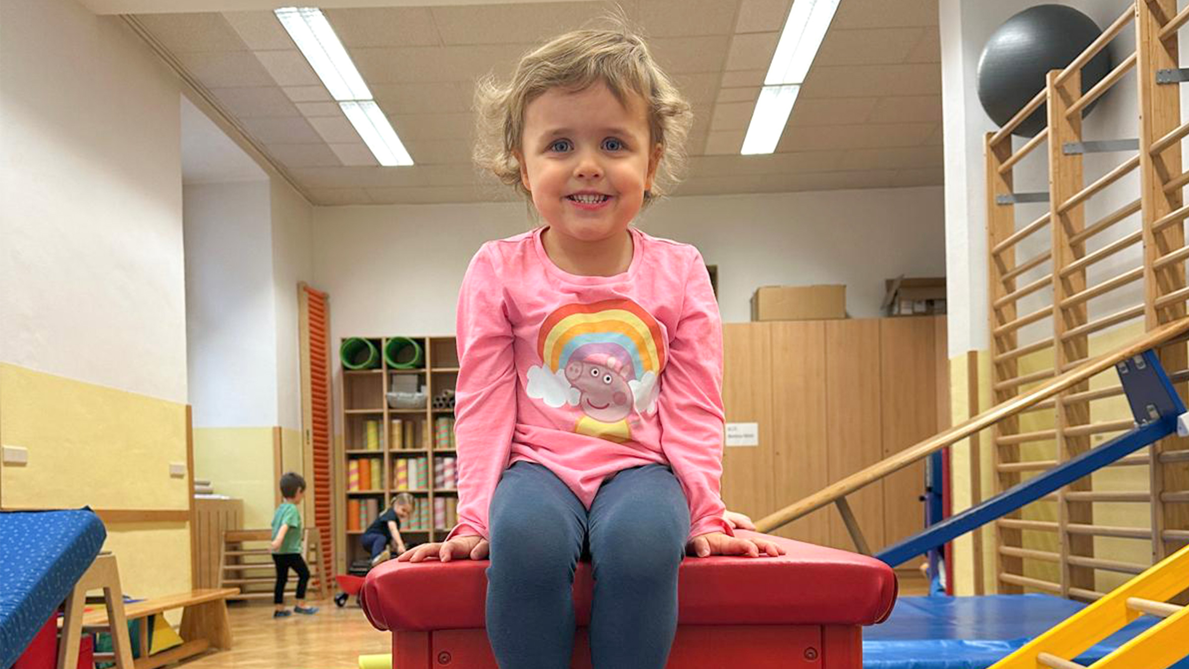 Kleines Mädchen mit rosarotem Shirt und blauer Legging auf einem Kasten in einem kleinen Turnsaal sitzend.