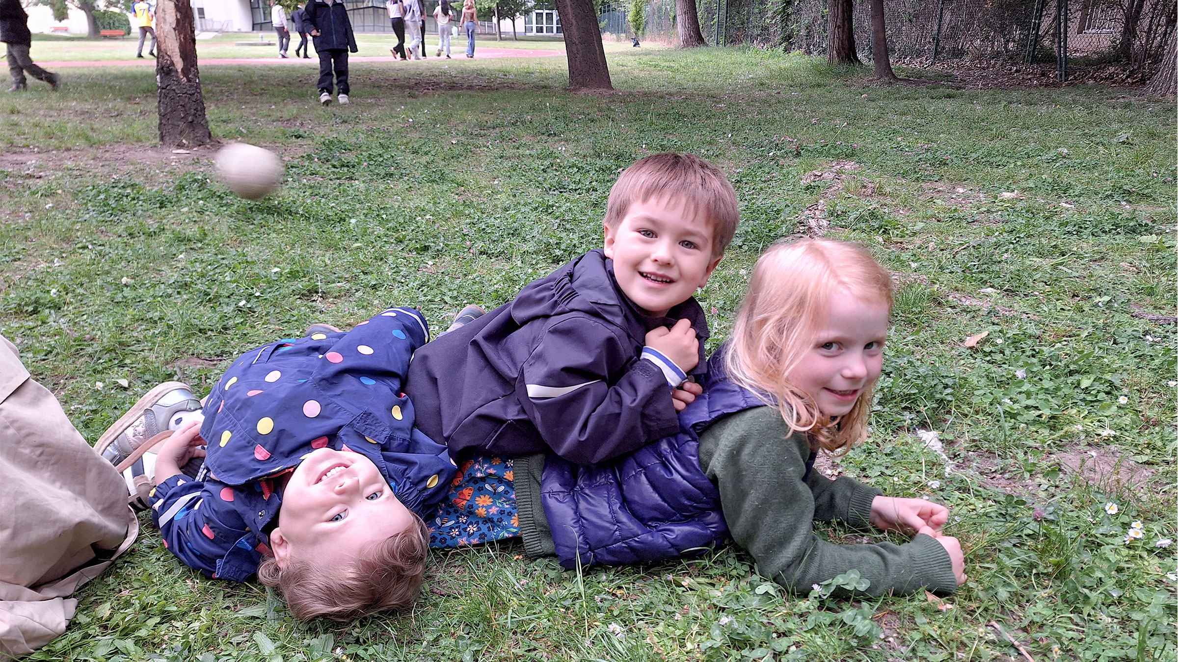 Drei Kinder mit Herbstkleidung auf der Wiese herumkugelnd und lachend in die Kamera blickend.