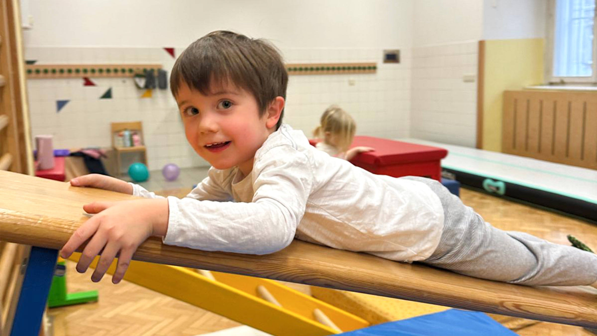 Kleiner Bub mit dunklen kurzen Haaren in die Kamera lachend auf einer Langbank sich nach oben ziehend