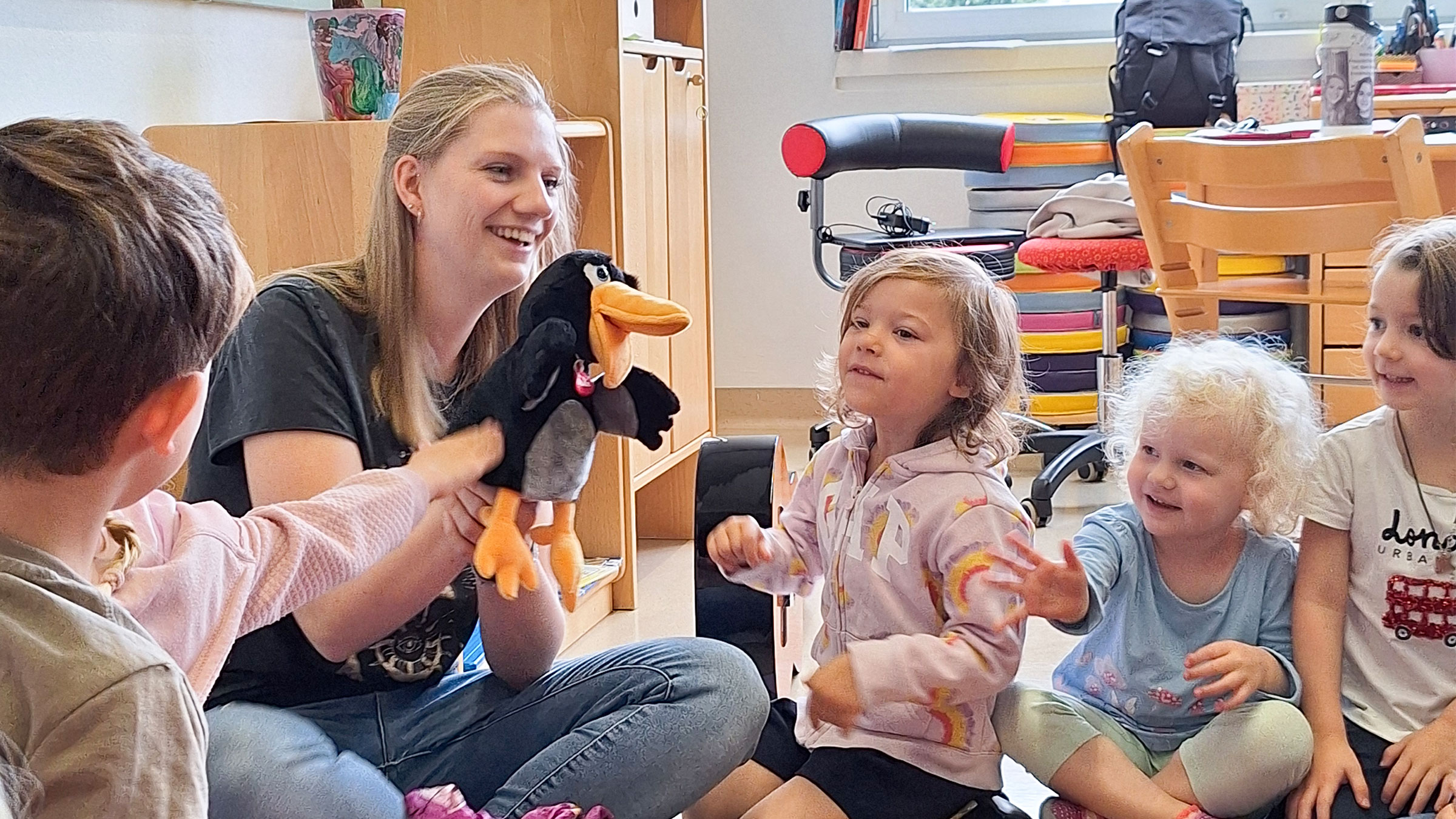 Lachende blonde Frau mit Handpuppe und mehrere lachende, neugierige Kinder am Boden um sie herum sitzend.