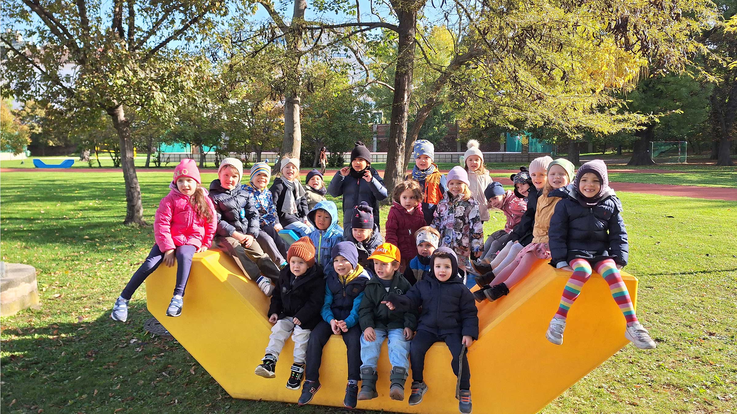 Gruppe von Kleinkindern mit Herbstkleidung in einem Park auf einem orangefarbenen Enzi sitzend und in die Kamera lachend