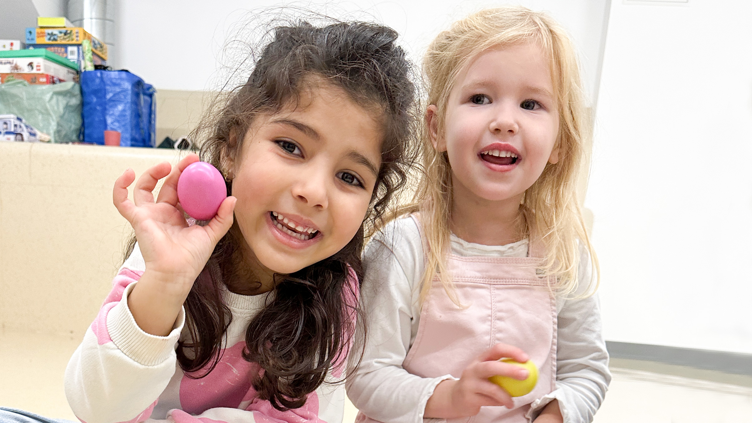 Zwei kleine Mädchen lachend in die Kamera blickend mit rosa und gelbem Rasselinstrument.