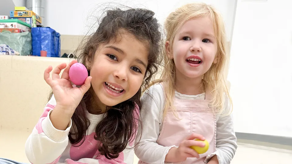 Zwei kleine Mädchen lachend in die Kamera blickend mit rosa und gelbem Rasselinstrument.