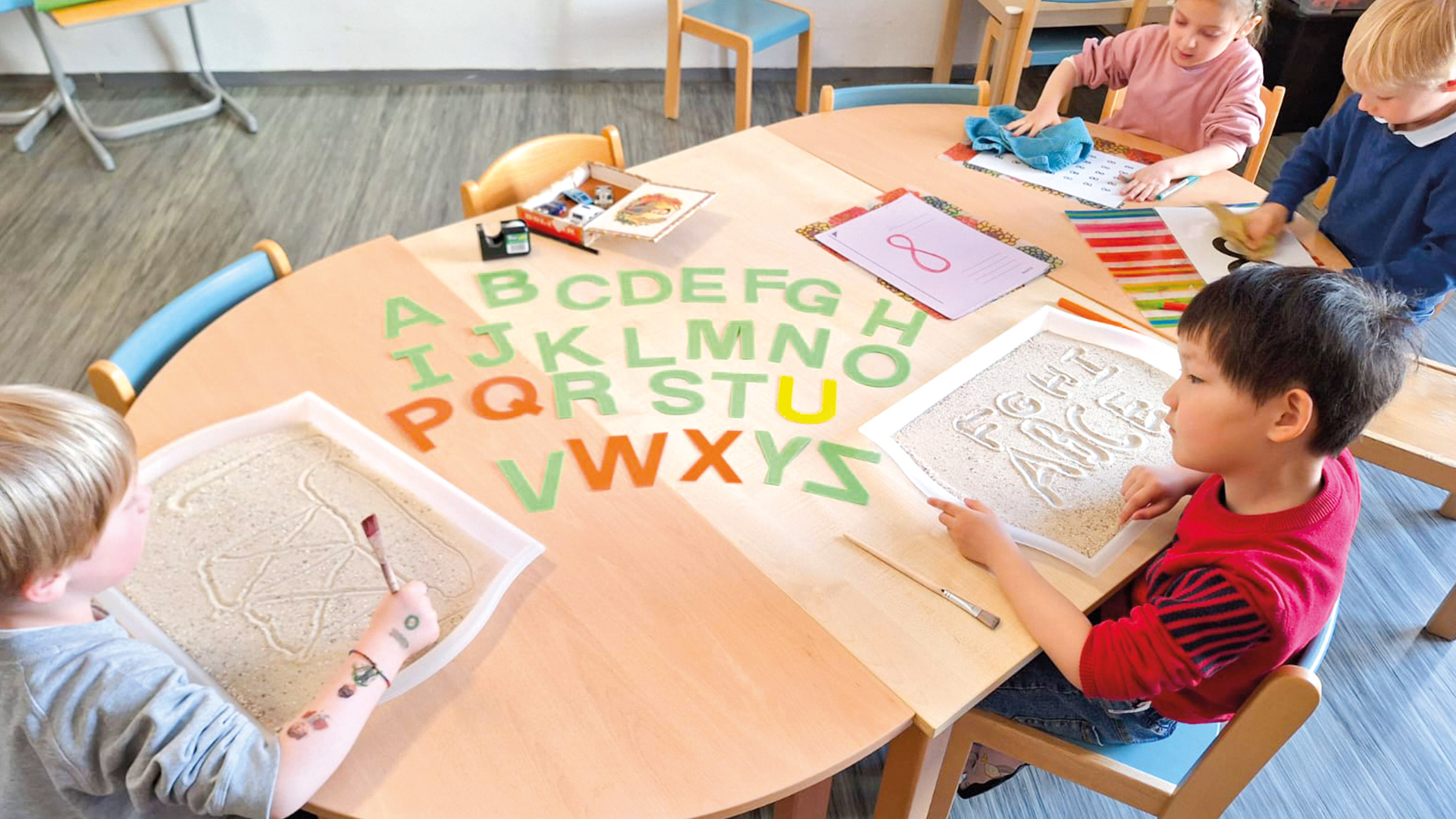 Tisch mit bunten Großbuchstaben und kleineren Kindern drumherum mit kleinen Sandboxen