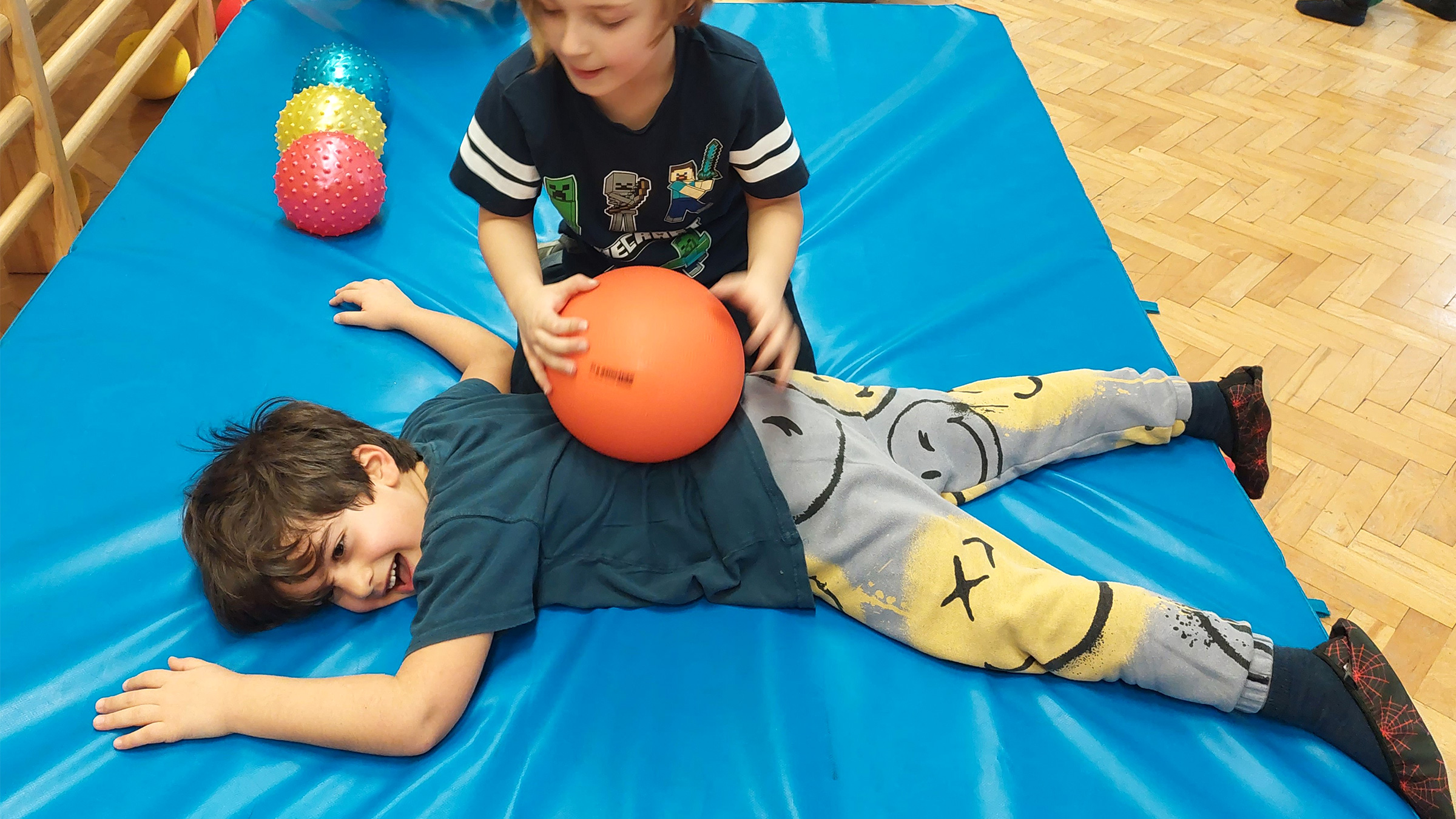 Bub am Bauch auf einer blauen Matratze liegend, alle Viere von sich streckend, mit lachendem Gesicht und einem Buben hinter ihm der einen orangenen Ball auf seinem Rücken rollt.