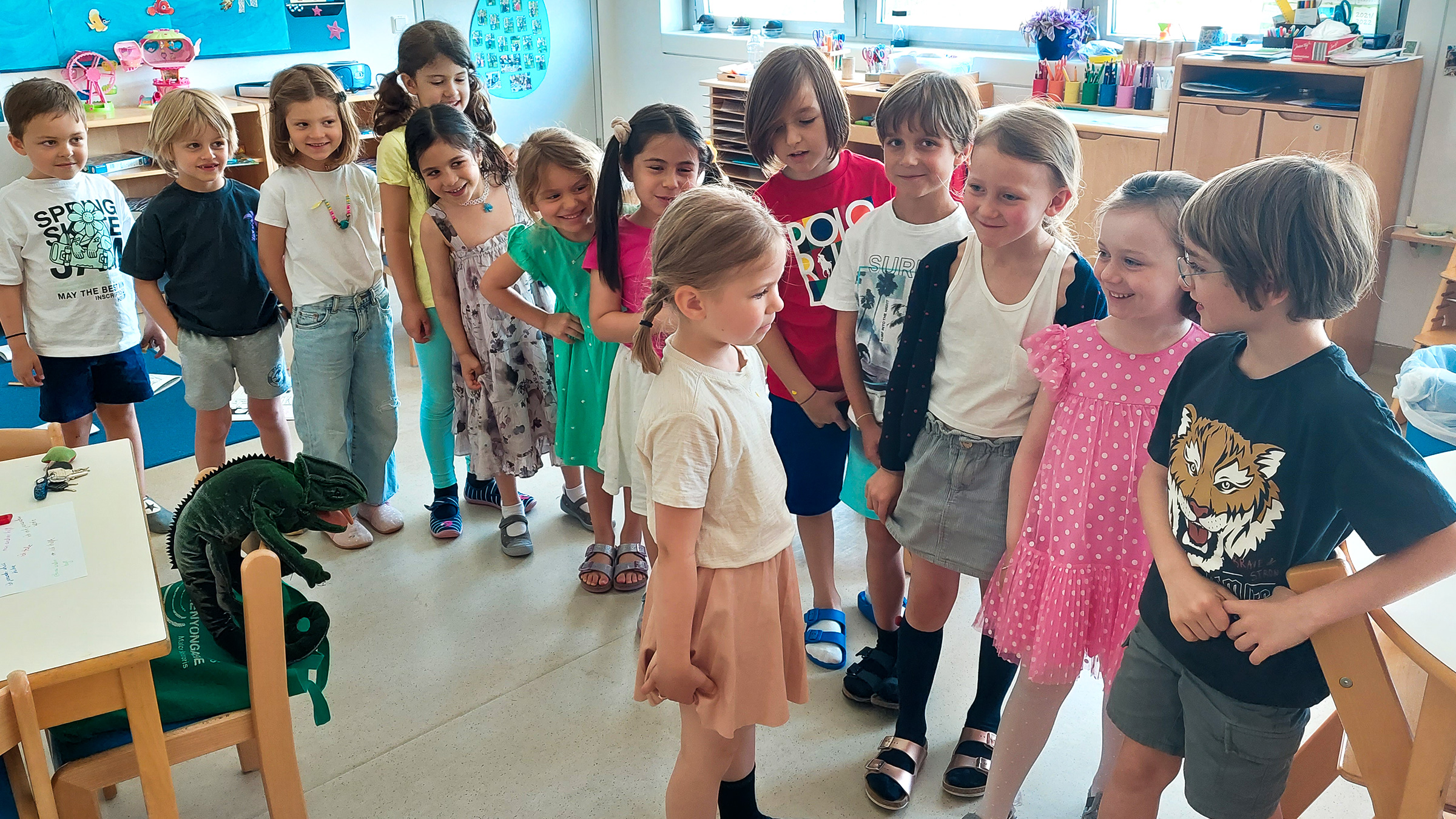 Gruppe von Kindern in Sommerkleidung in einer Reihe stehend und gespannt auf ein schüchtern wirkendes Mädchen blickend..