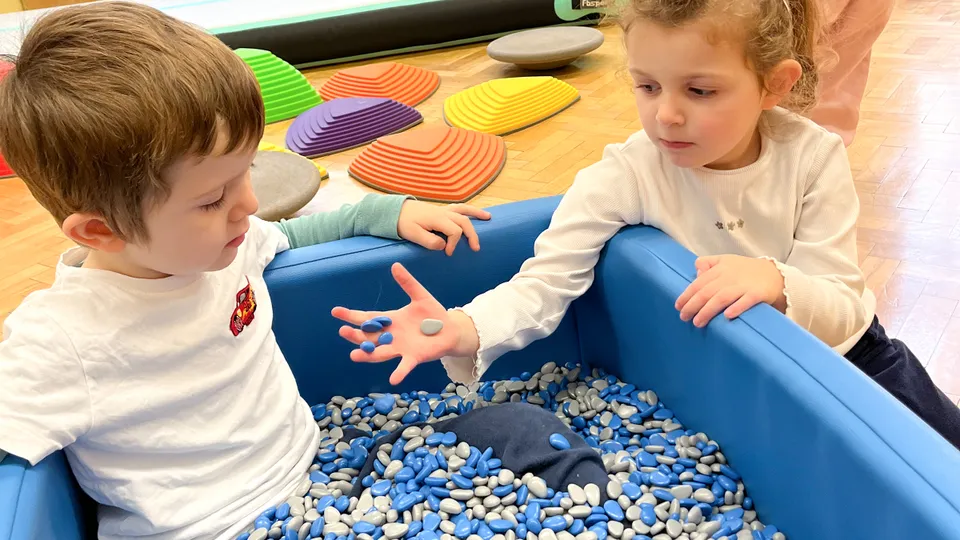 Ein Bub in einer blauen Kiste mit blau weißen Steinen sitzend. Daneben ein Mädchen das in sich gekehrt ein paar der Steine durch ihre Finger rieseln lässt.
