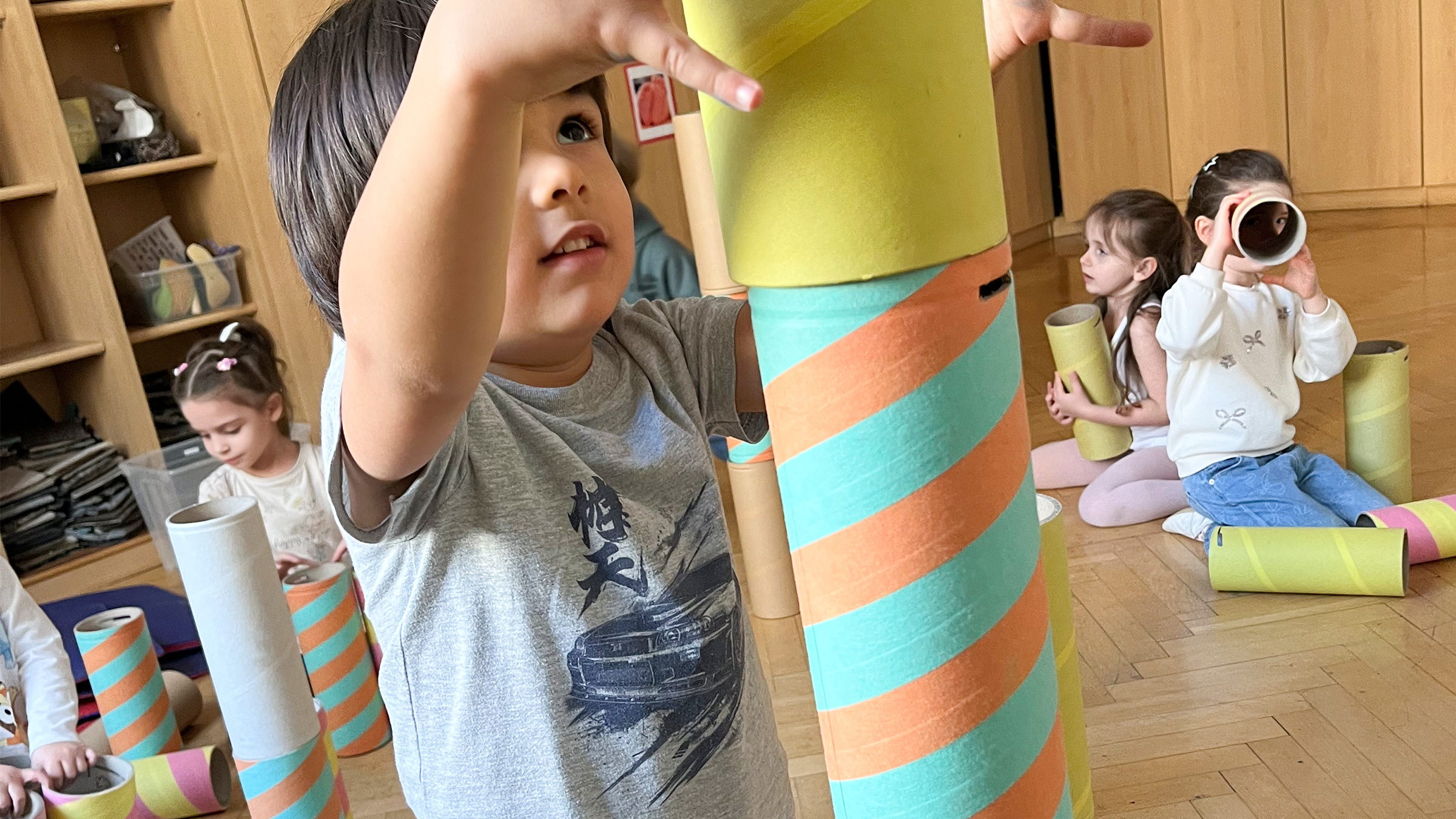 Nahaufnahme eines Buben in voller Konzentration beim Stapeln von bunten Kartonröhren mit weiteren Kindern im Hintergrund, die mit ähnlichen Röhren spielen.