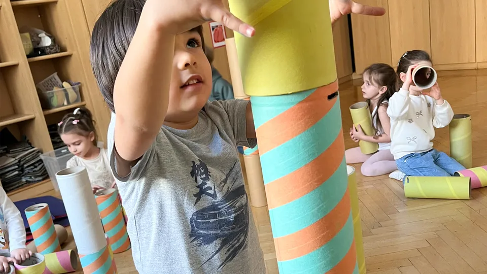 Nahaufnahme eines Buben in voller Konzentration beim Stapeln von bunten Kartonröhren mit weiteren Kindern im Hintergrund, die mit ähnlichen Röhren spielen.