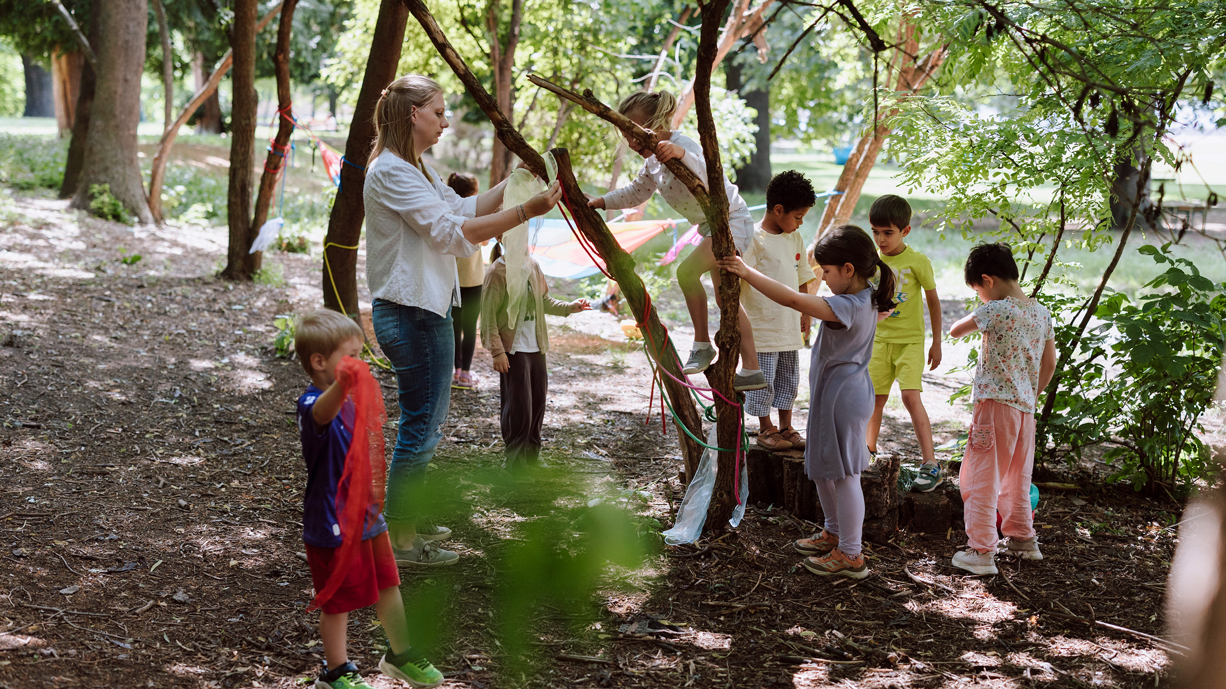 Gruppe von Kindern in Sommerkleidung mit einer erwachsenen Frau in einem lichten Wald mit bunten Tüchern spielend und auf Baumstämme kletternd