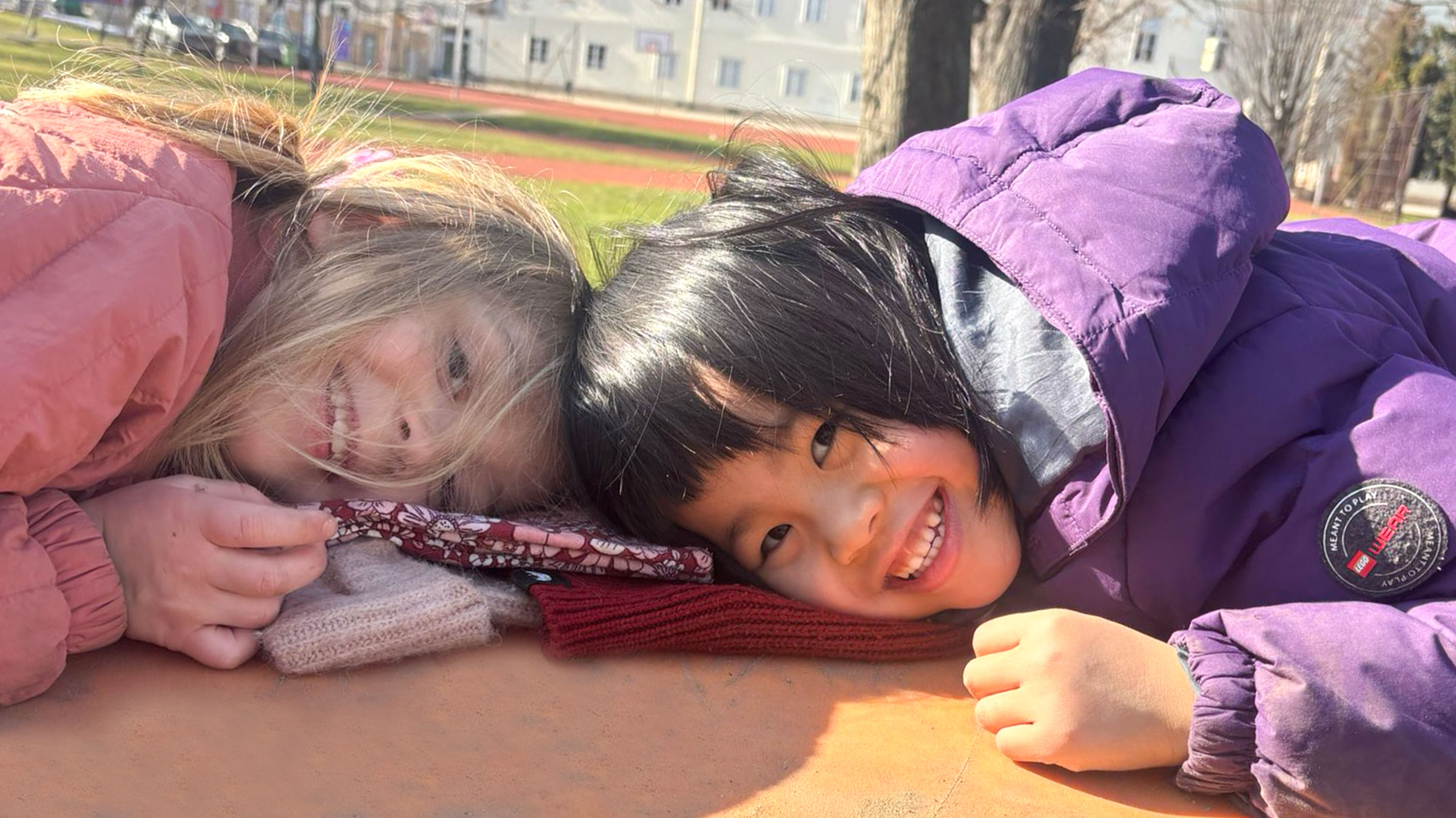 Zwei Mädchen in warmer Kleidung bei sonnigem Wetter mit dem Oberkörper auf einer orangenen Plattform liegend, die Köpfe zusammensteckend, in die Kamera lachend