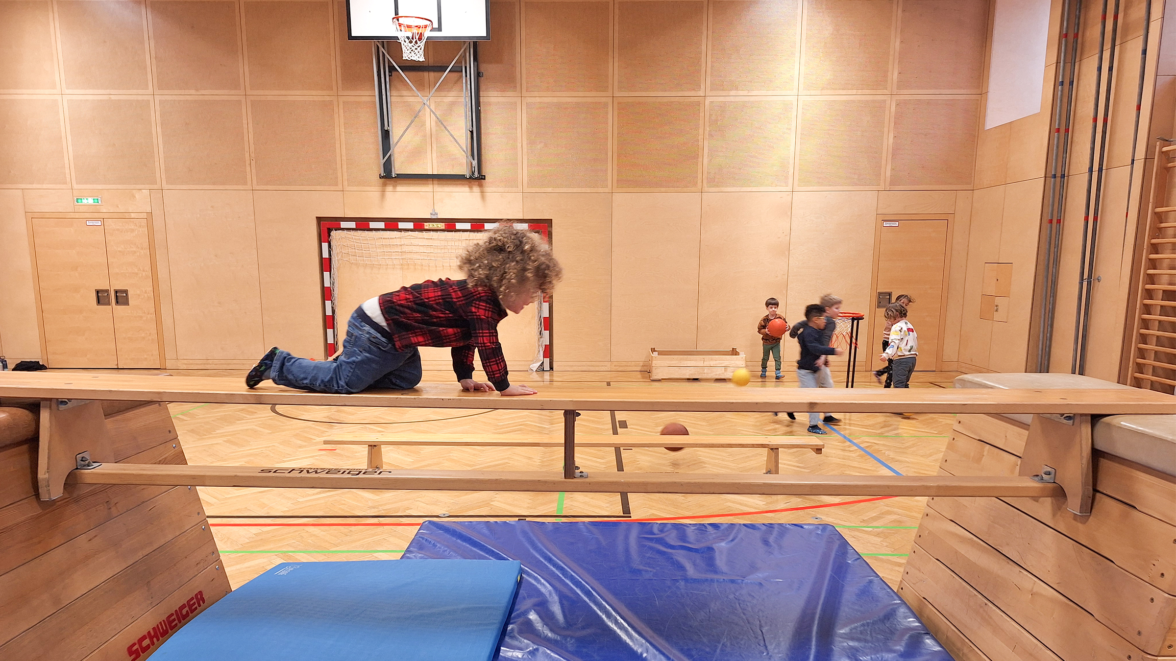 Kleinkind über eine Langbank auf allen Vieren klettern in einem großen Turnsaal mit blauem Weichboden darunter
