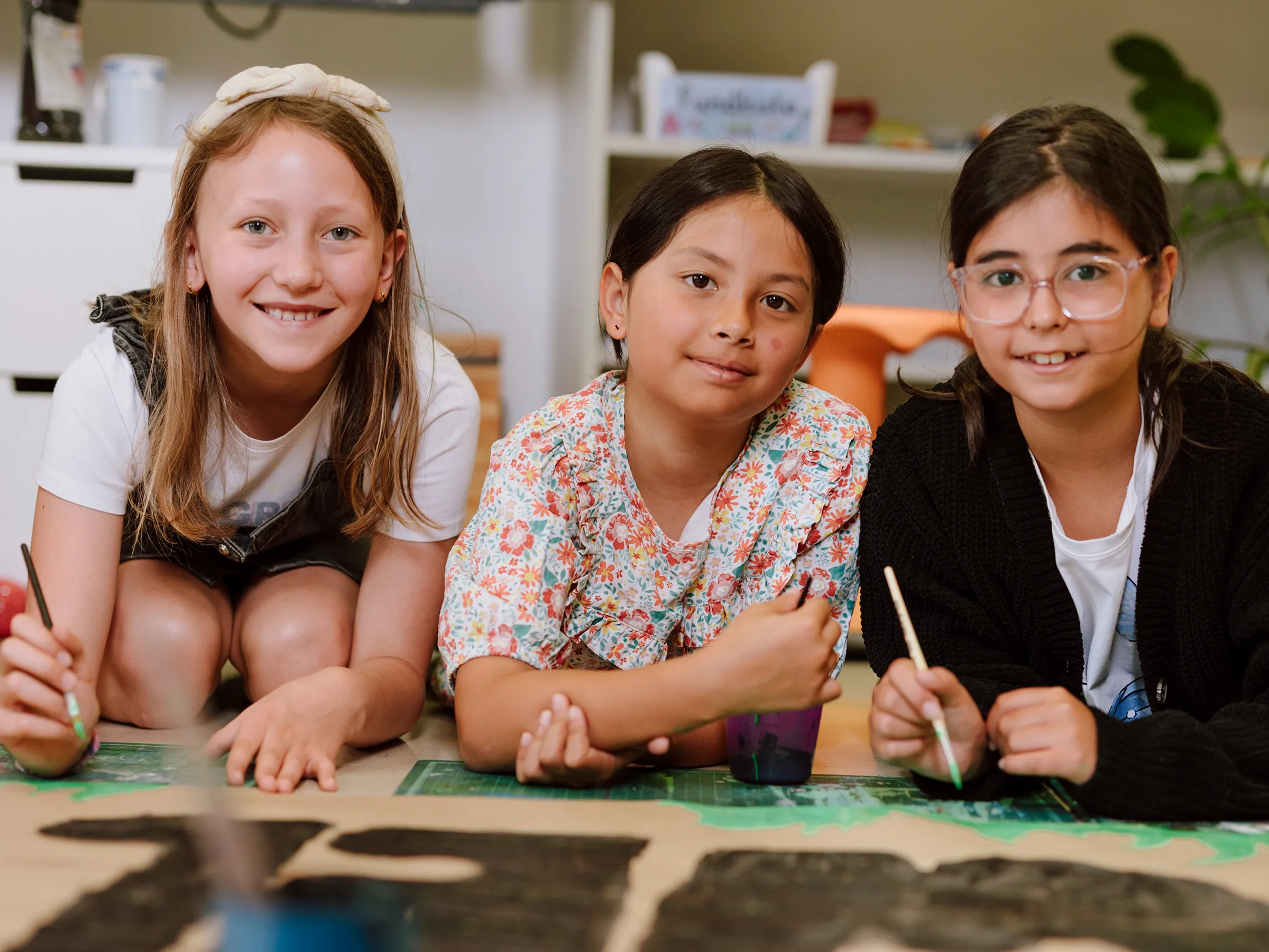 Drei junge Mädchen am Boden liegend bzw. knieend in die Kamera lachend während sie an einem plakat mit wasserfarben arbeiten