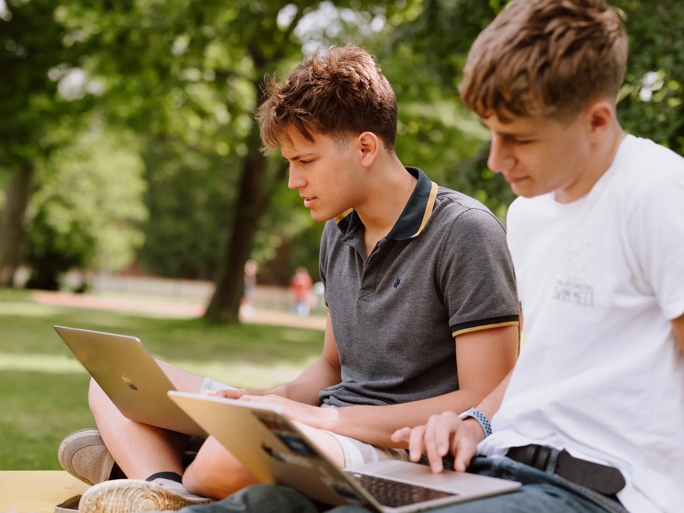 Zwei junge Männer in einem Park gemütlich mit Laptop auf dem Schoss fokussiert auf die Bildschirme blickend