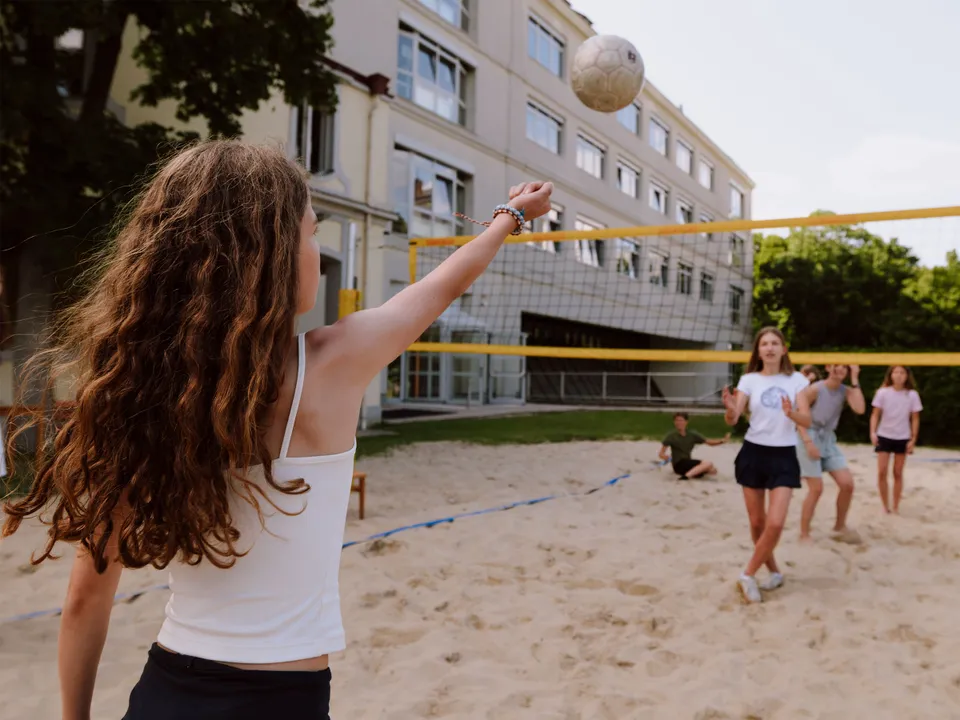 Mädchen mit langen gewellten Haaren und weißem Top einen Ball in Richtung Beachvolleyballnetz spielend.