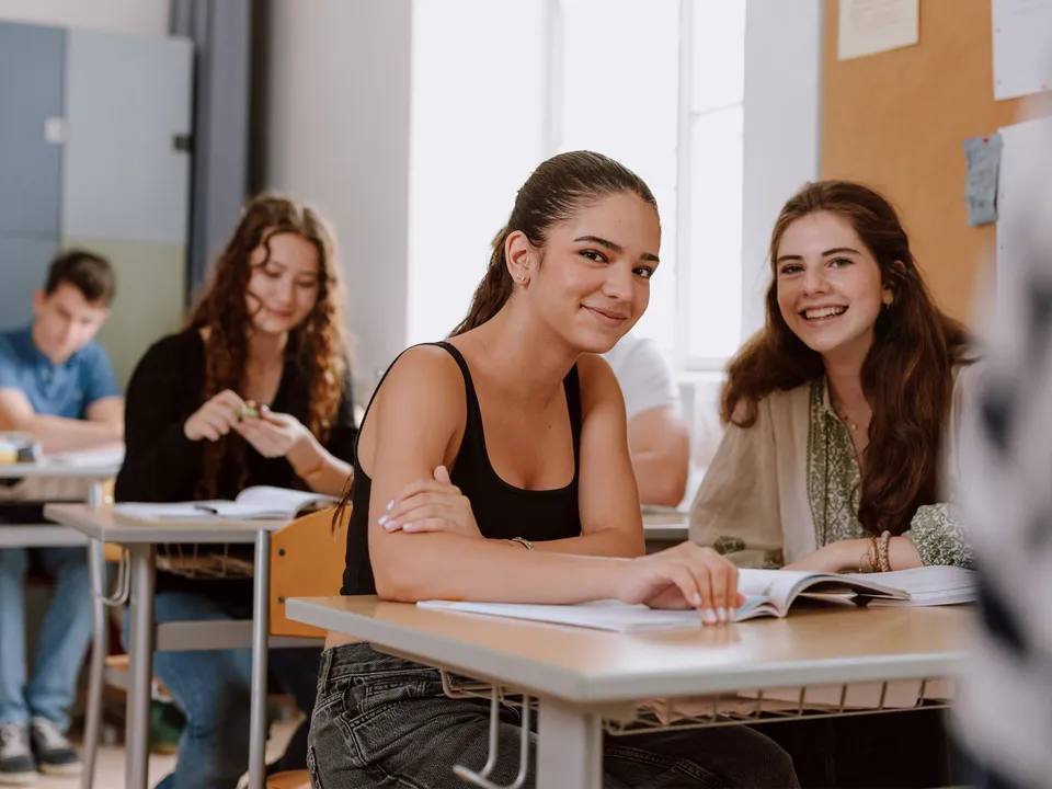 Zwei weibliche Teenager mit langen dunklen Haaren in sommerlicher Kleidung in einem Klassenraum sitzend und lachend in die Kamera blickend