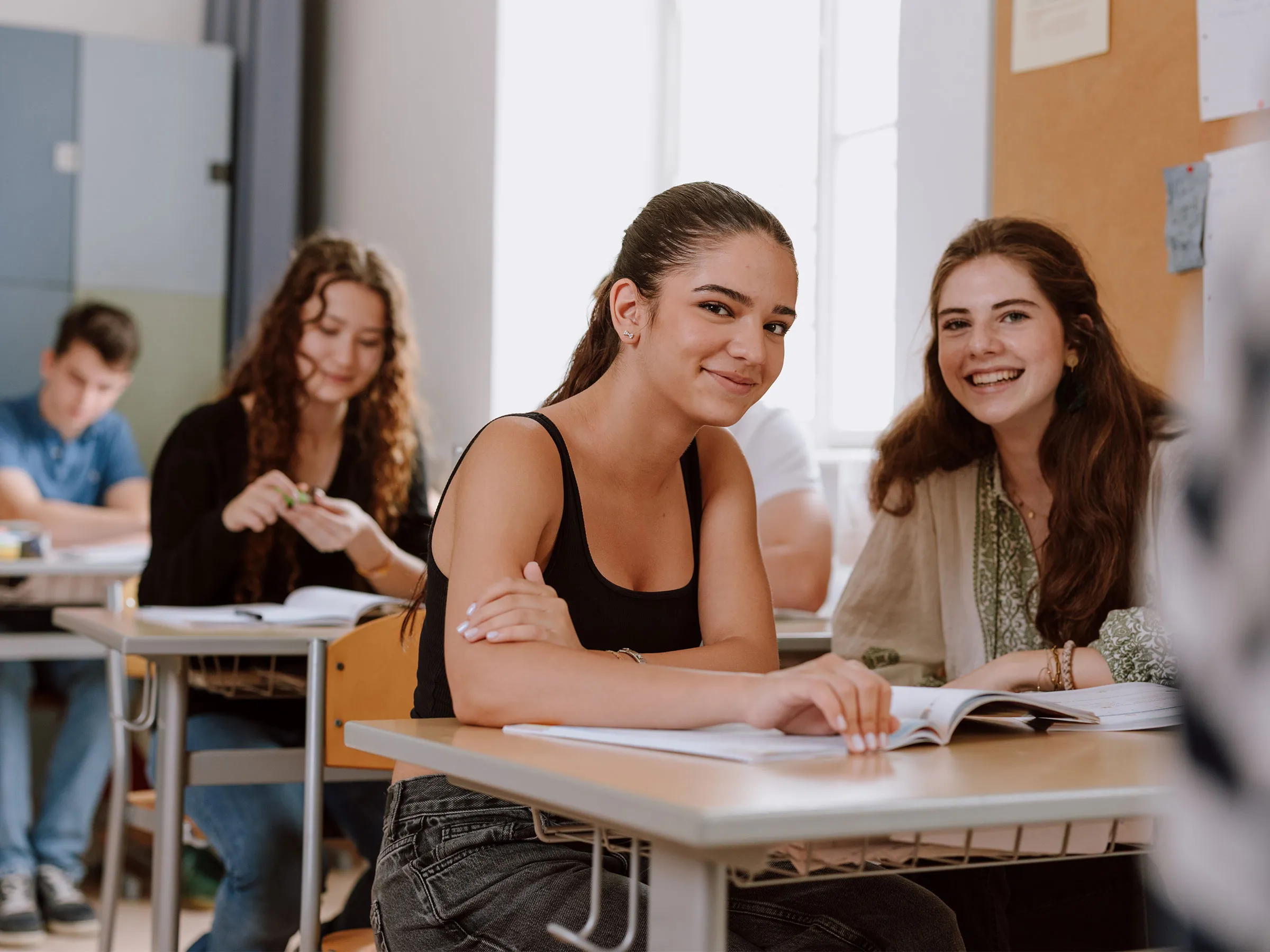 Zwei weibliche Teenager mit langen dunklen Haaren in sommerlicher Kleidung in einem Klassenraum sitzend und lachend in die Kamera blickend