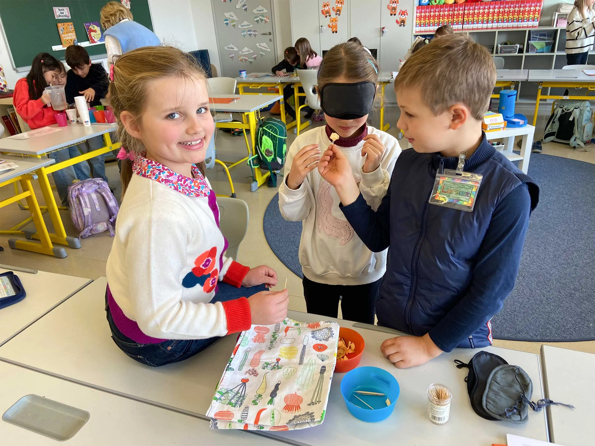 3 Kinder in einem Klassenzimmer bei einem Tisch stehend. Ein Mädchen hat eine Augenbinde und bekommt ein Stück Essen auf einem Zahnstocher gereicht.