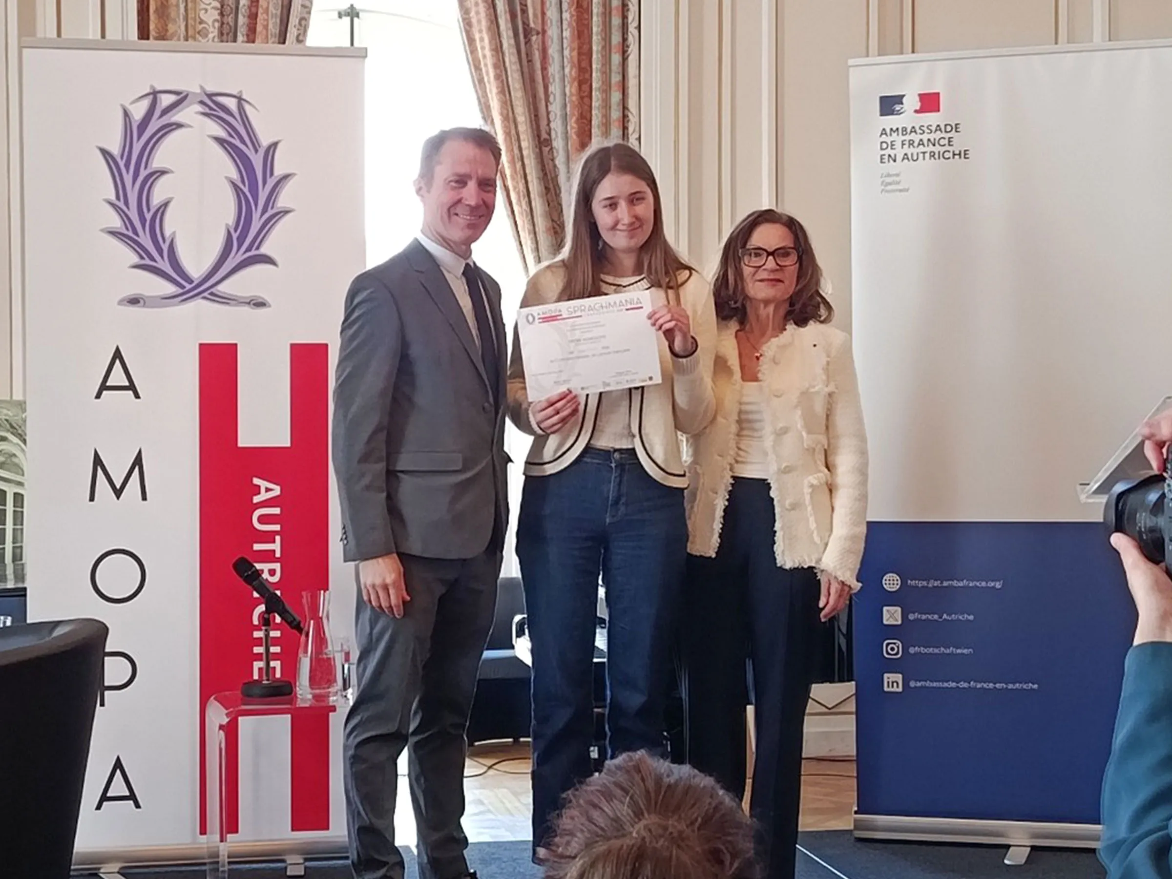 Eine junge Frau, flankiert von einem Mann und einer Frau hält ein Zertifikat in die Kamera, mit Rollups der französischen Botschaft daneben.