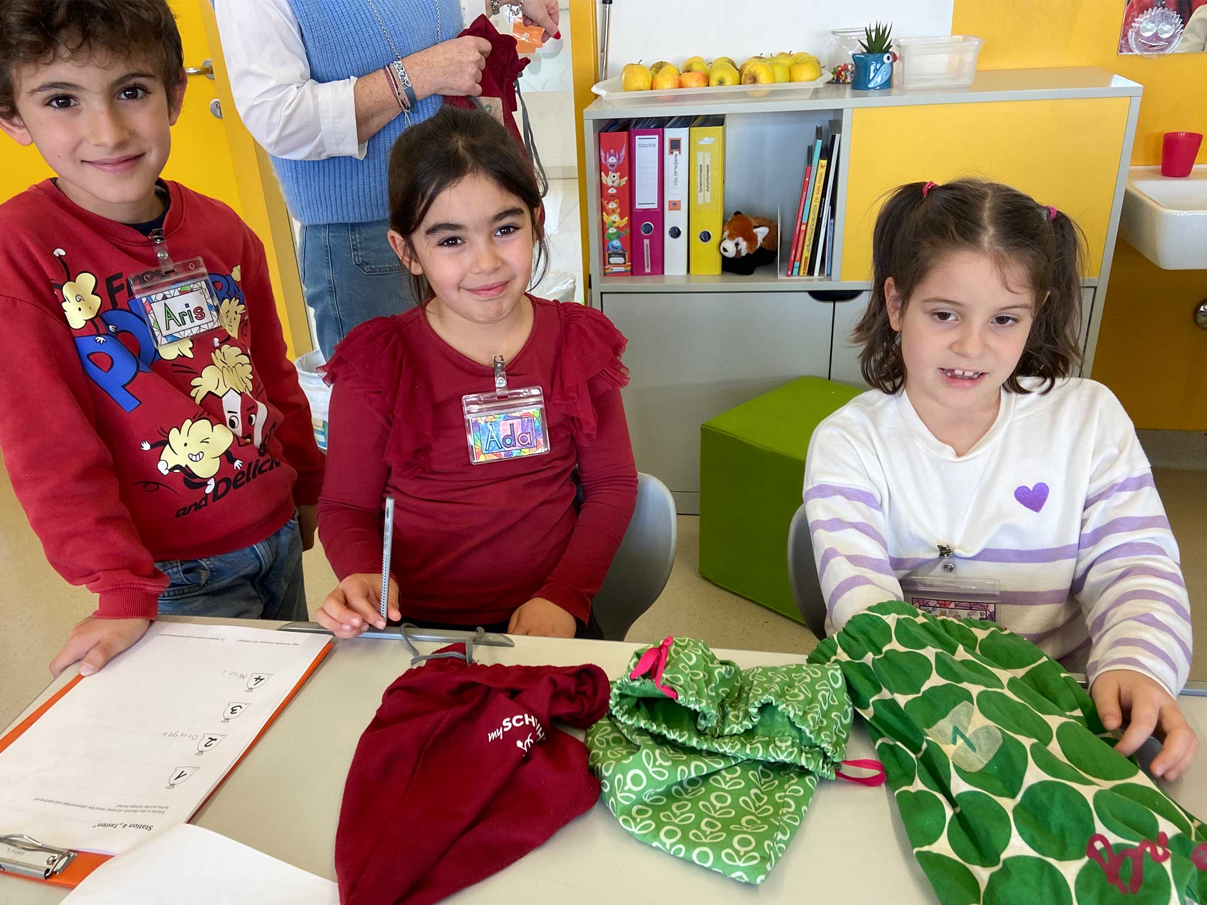 Drei Kinder an einem Tisch stehend mit bunten Stoffsäckchen vor sich liegend. Ein Kind greift in eines der Säckchen hinein.
