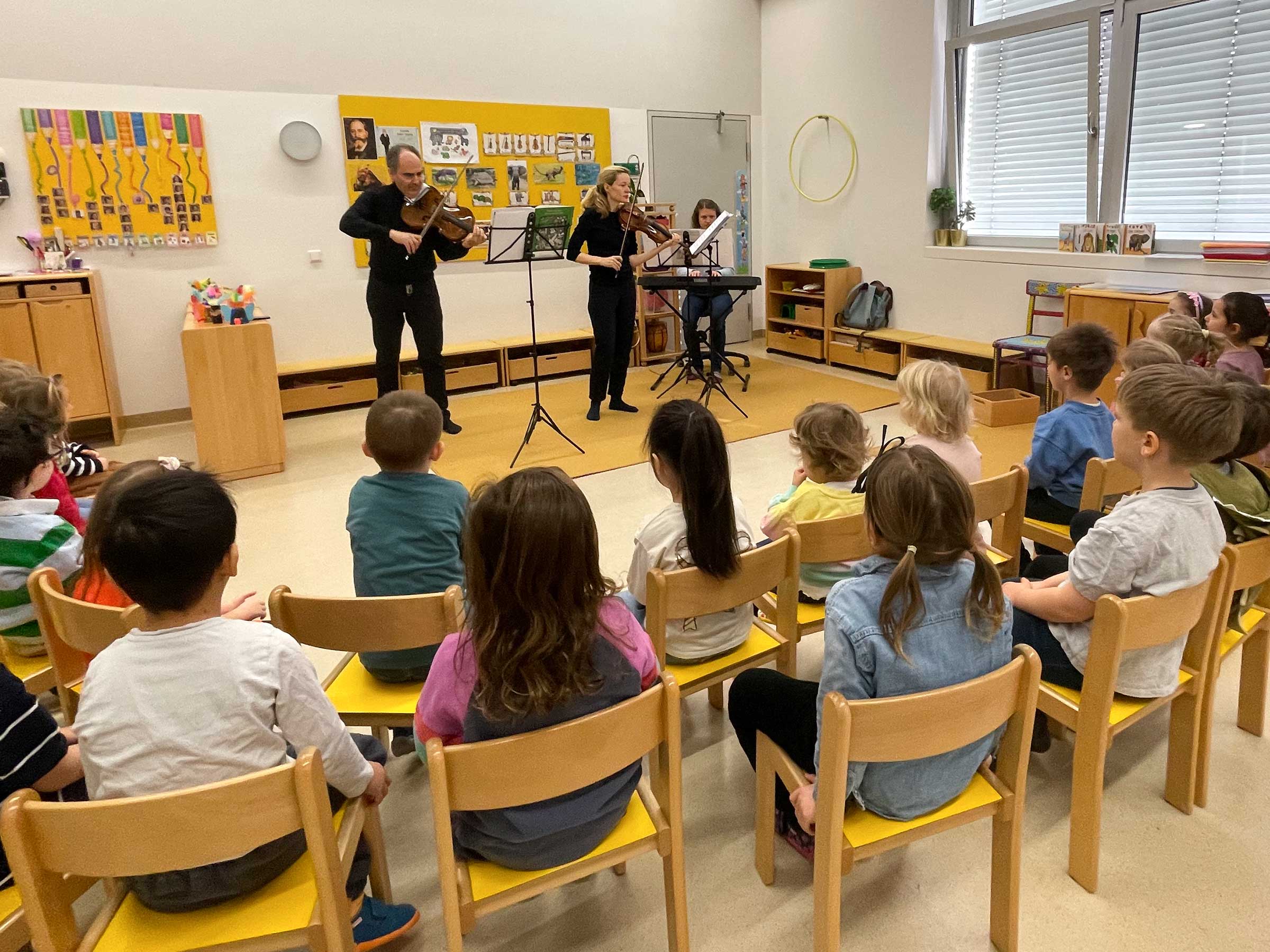Kinder in einem Stuhlkreis um zwei erwachsene Personen mit Geige sitzend.