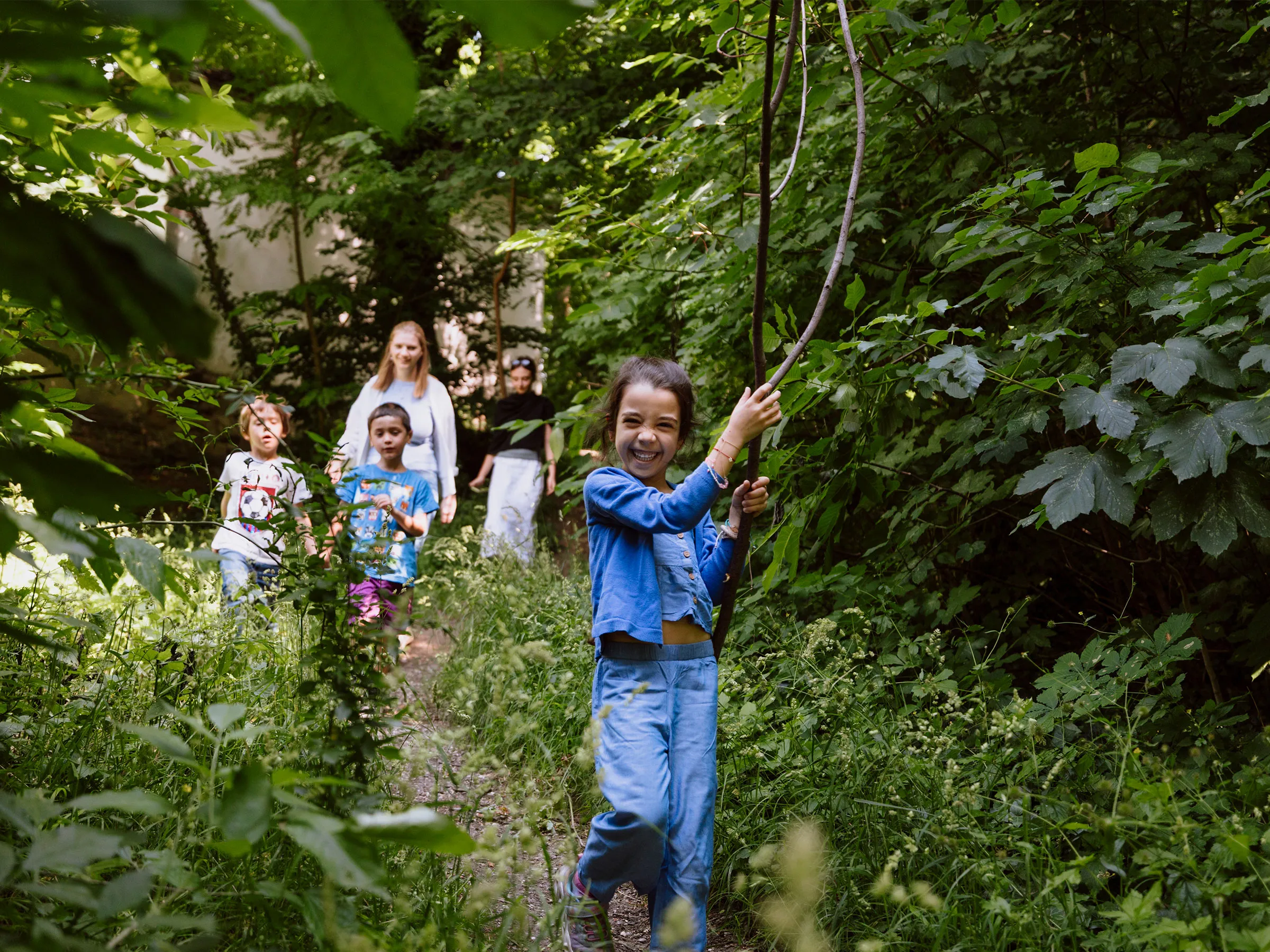Gruppe von Kindern in einem Waldstück an einem sonnigen Sommertag mit einem lachenden Mädchen vorausgehend und einen großen Ast tragend.